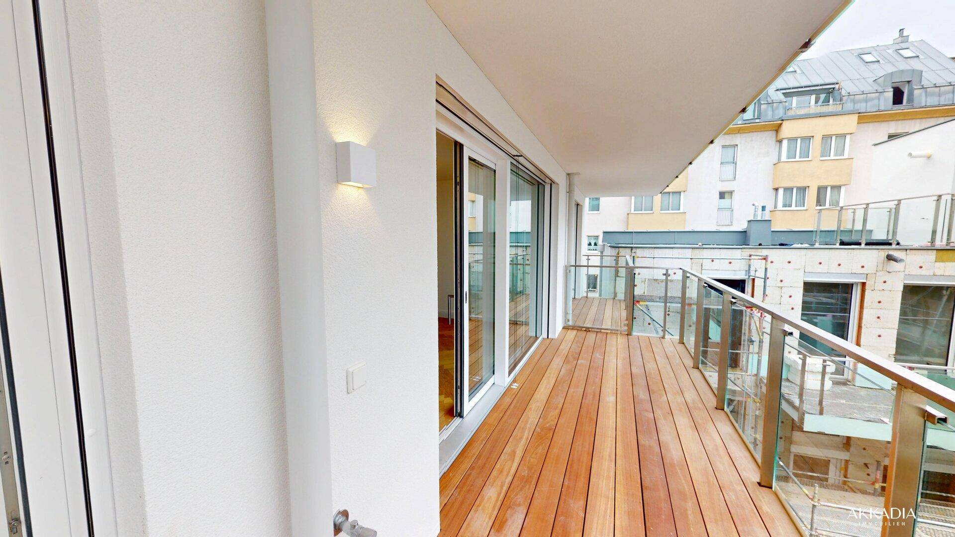 Heller Balkon mit Holzbelag, Glasgeländer und Blick auf die umliegenden Gebäude der Stadt.