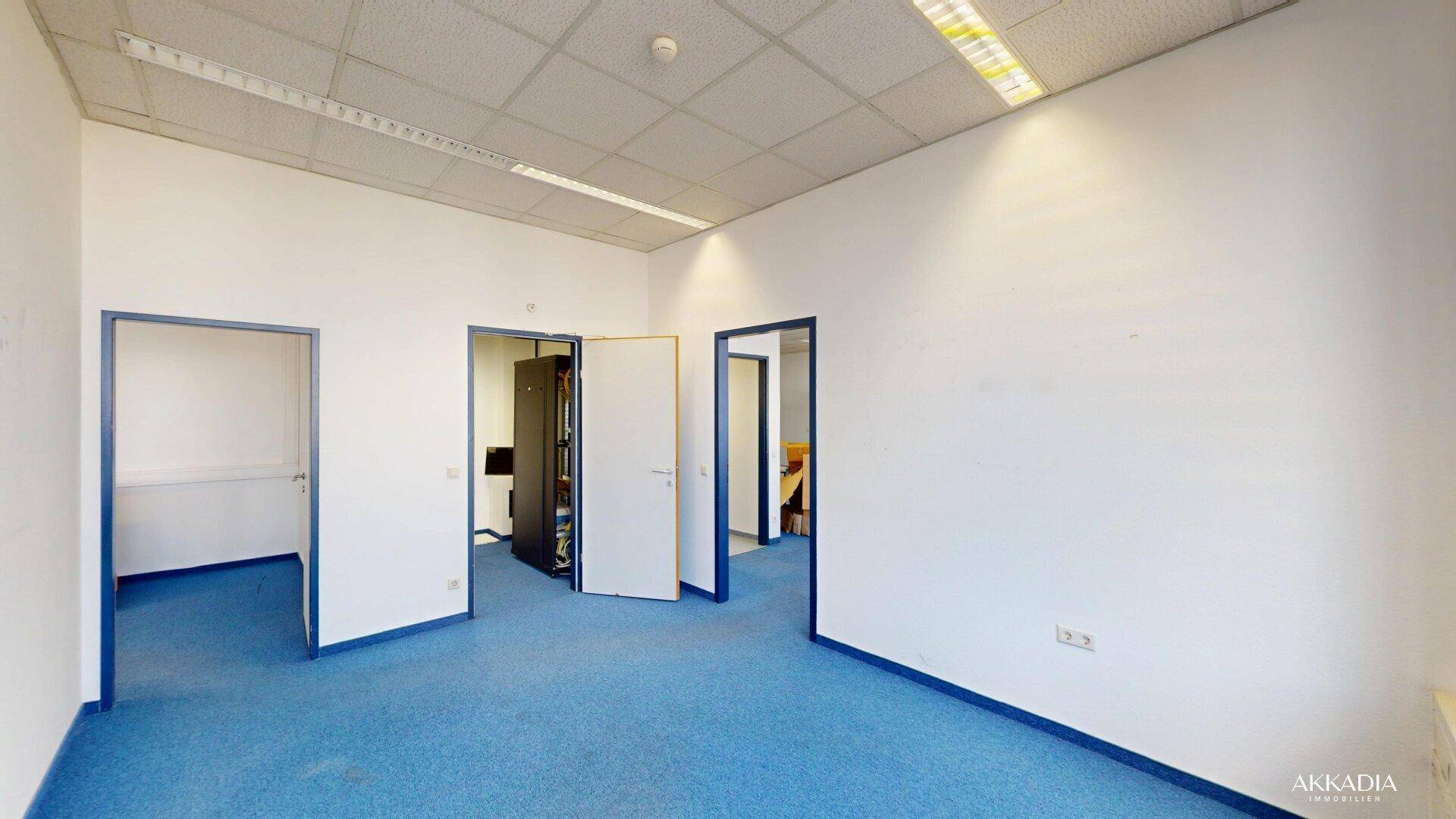 Leerer Büroraum mit blauem Teppichboden und mehreren Türen, die zu angrenzenden Räumen führen.