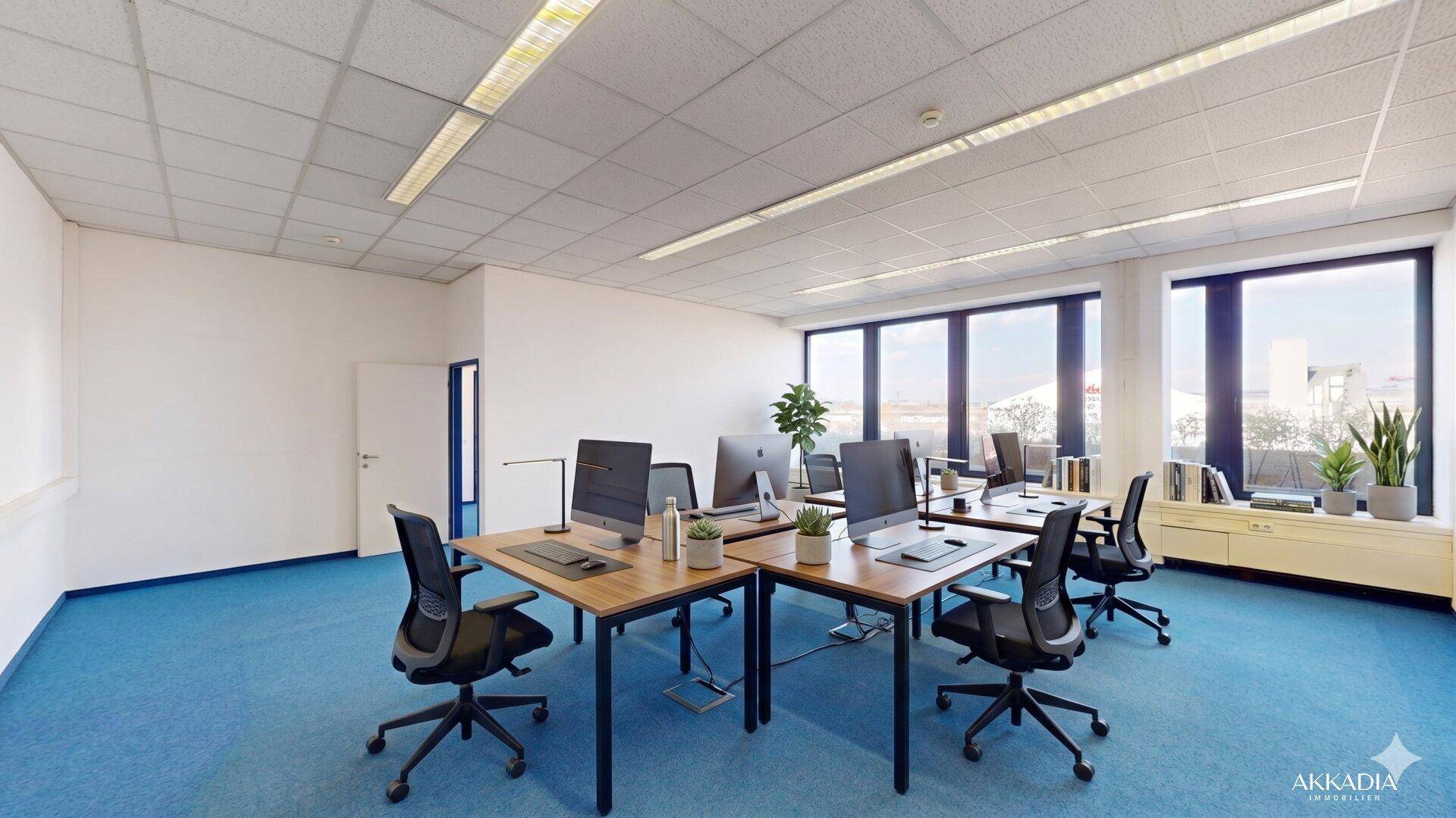 Modernes Büro mit mehreren Arbeitsplätzen, blauer Teppichboden und großen Fenstern mit Ausblick.