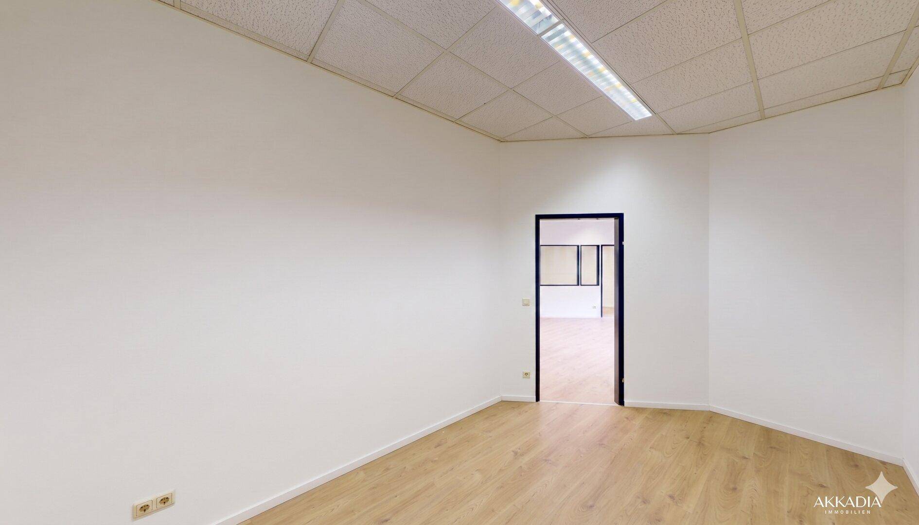 Leerer Büroraum mit hellem Laminatboden und einer Tür, die zu einem weiteren Bereich führt, gut beleuchtet.
