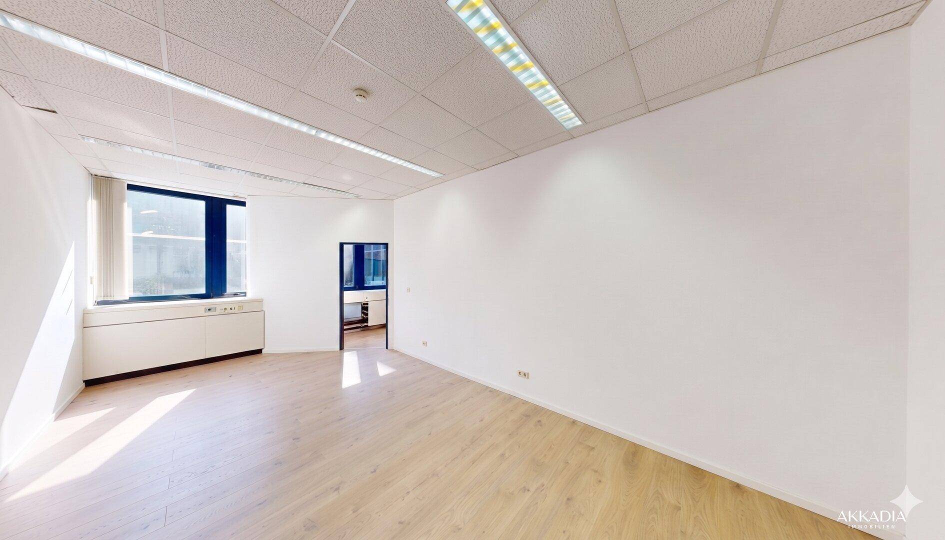 Leerer Büroraum mit Holzboden und einem großen Fenster, das viel Tageslicht spendet.
