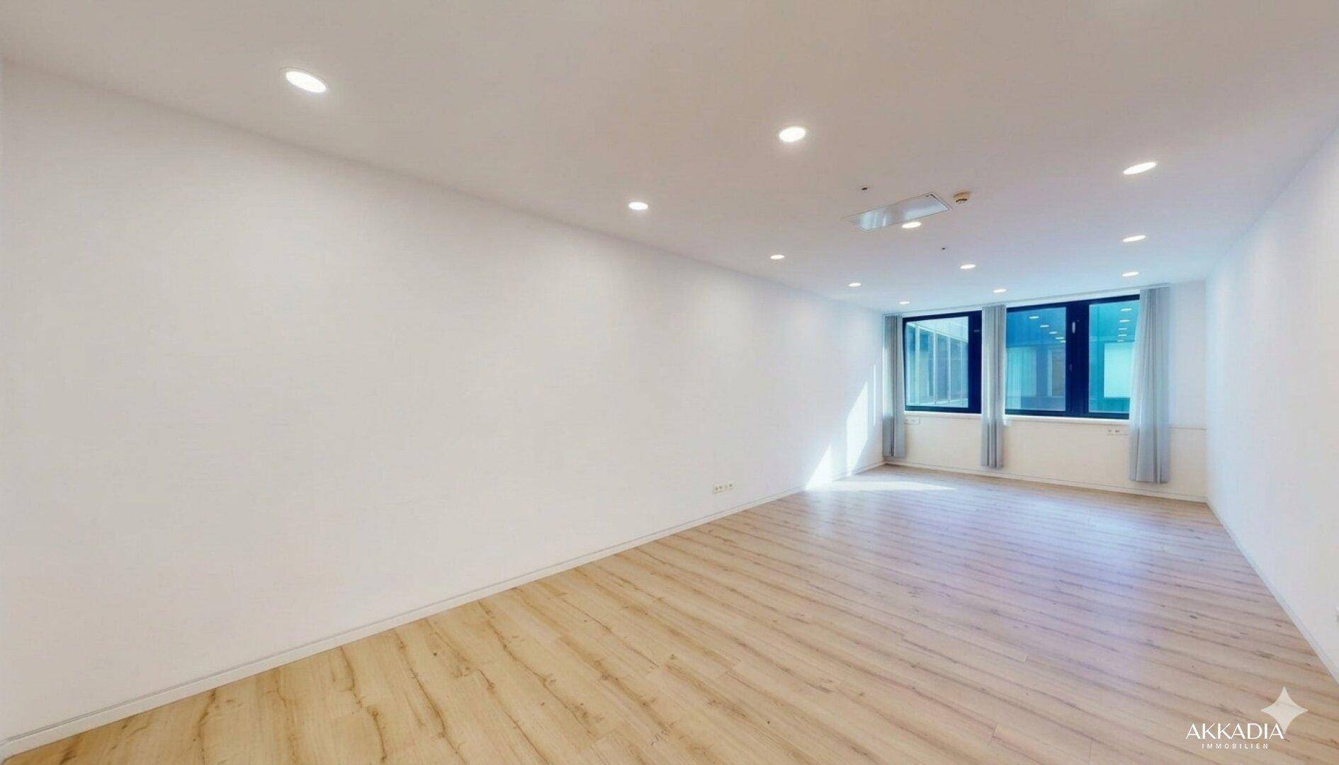 Heller Büroraum mit großen Fenstern, die viel Tageslicht hereinlassen, und modernem Laminatboden.