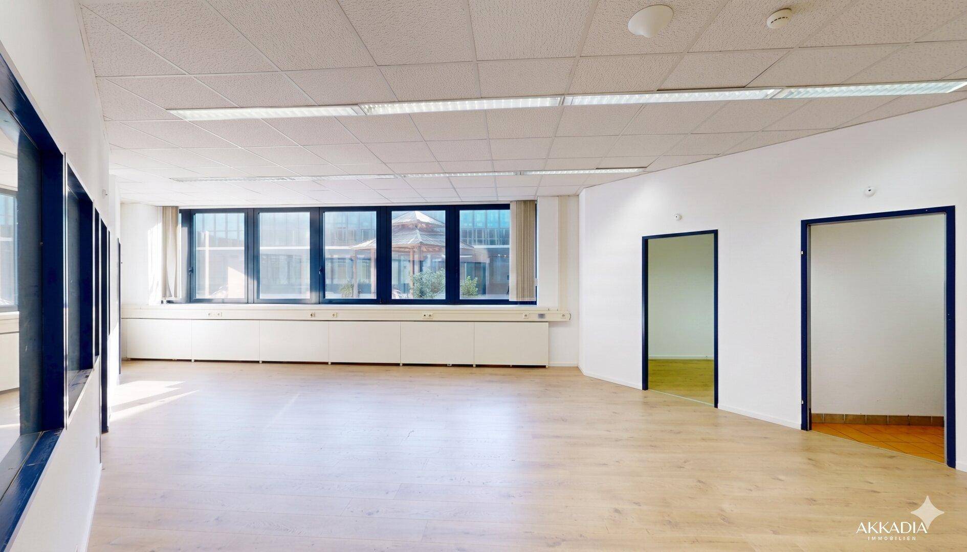 Leerer Büroraum mit Holzboden, Fenstern und Türen zu weiteren Bereichen.