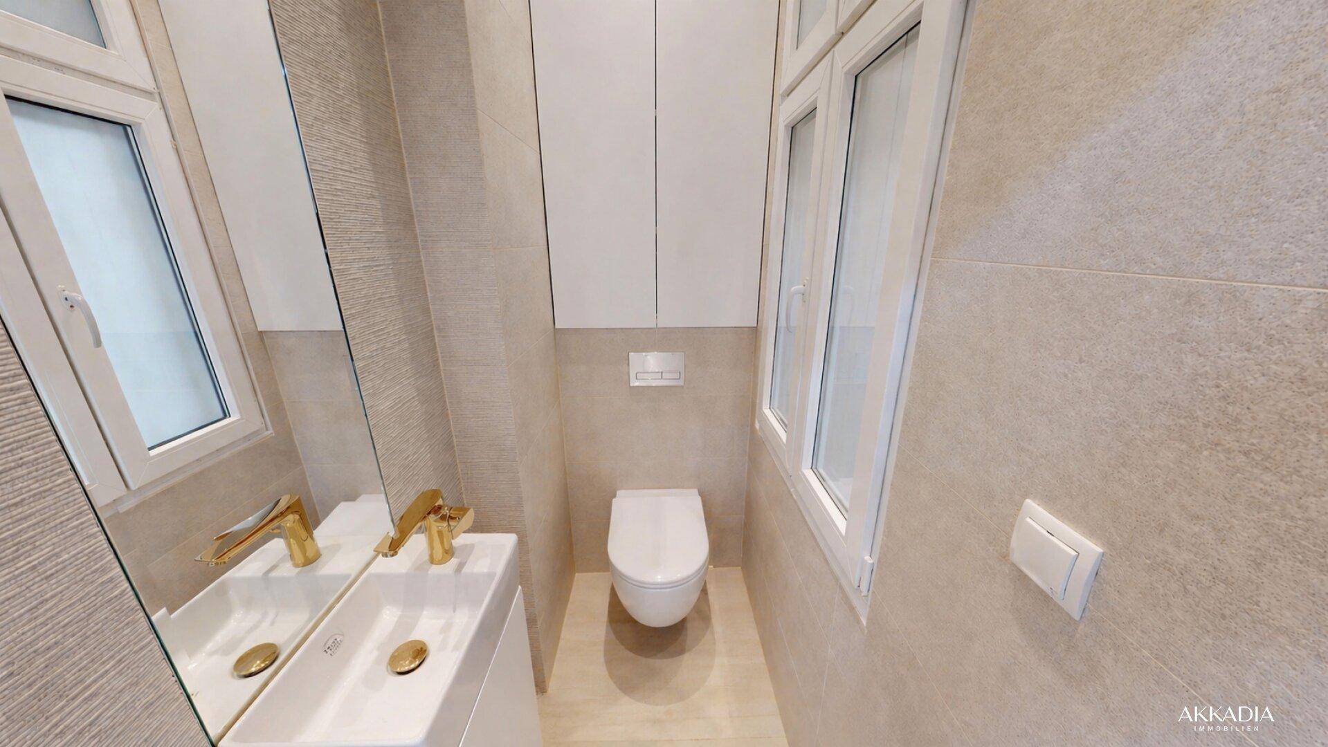 Modernes Gäste-WC mit wandmontierter Toilette, Fenster und goldenen Armaturen in beigefarbenen Fliesen.