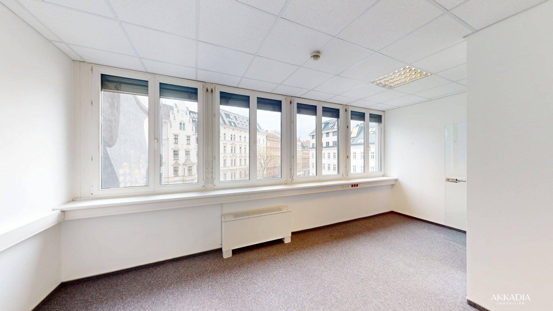 Heller Büroraum mit Teppichboden, weißen Wänden und einer langen Fensterfront mit Stadtblick.