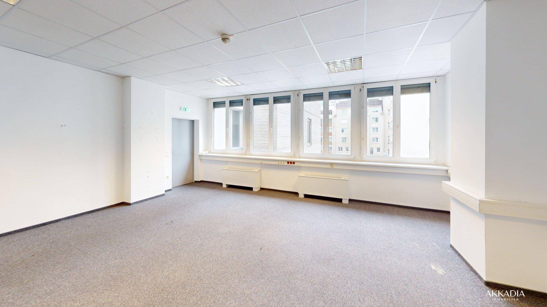 Geräumiger Büroraum mit Teppichboden, weißen Wänden und einer langen Fensterfront mit Stadtblick.