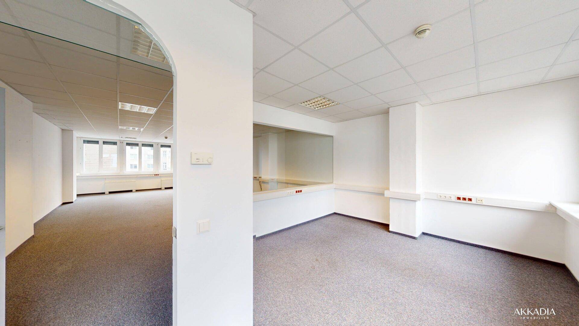 Geräumiges Büro mit Teppichboden und Blick in einen weiteren Bürobereich durch eine Glaswand.