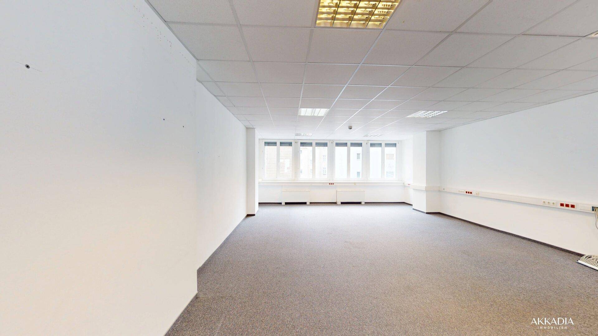 Großer, leerer Büroraum mit Teppichboden und einer Fensterfront, die den Raum hell erscheinen lässt.