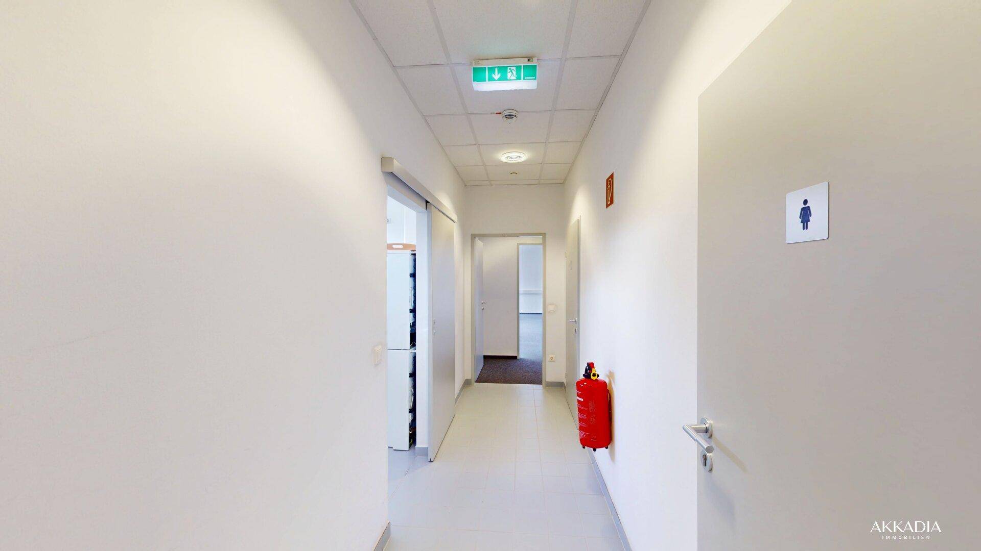 Heller Flur mit weißen Wänden, Notausgangsschild und Feuerlöscher, der zu weiteren Räumen führt.