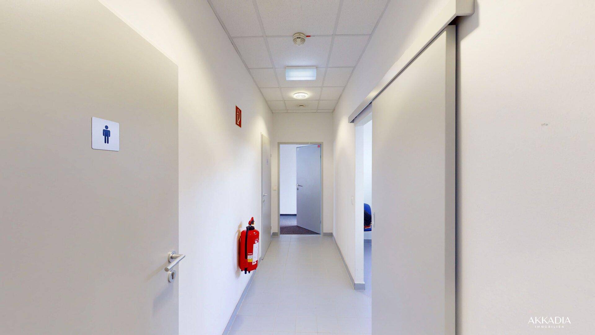 Schmaler Flur mit weißen Wänden, Feuerlöscher und Türen zu den Toilettenbereichen.