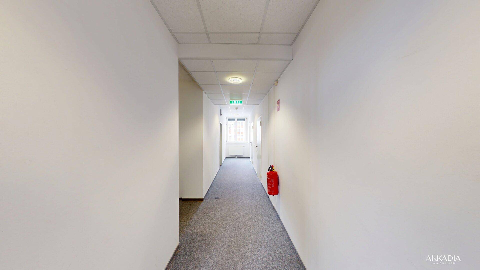Langer, heller Flur mit Teppichboden und weißen Wänden, der zu einem Fenster am Ende führt.