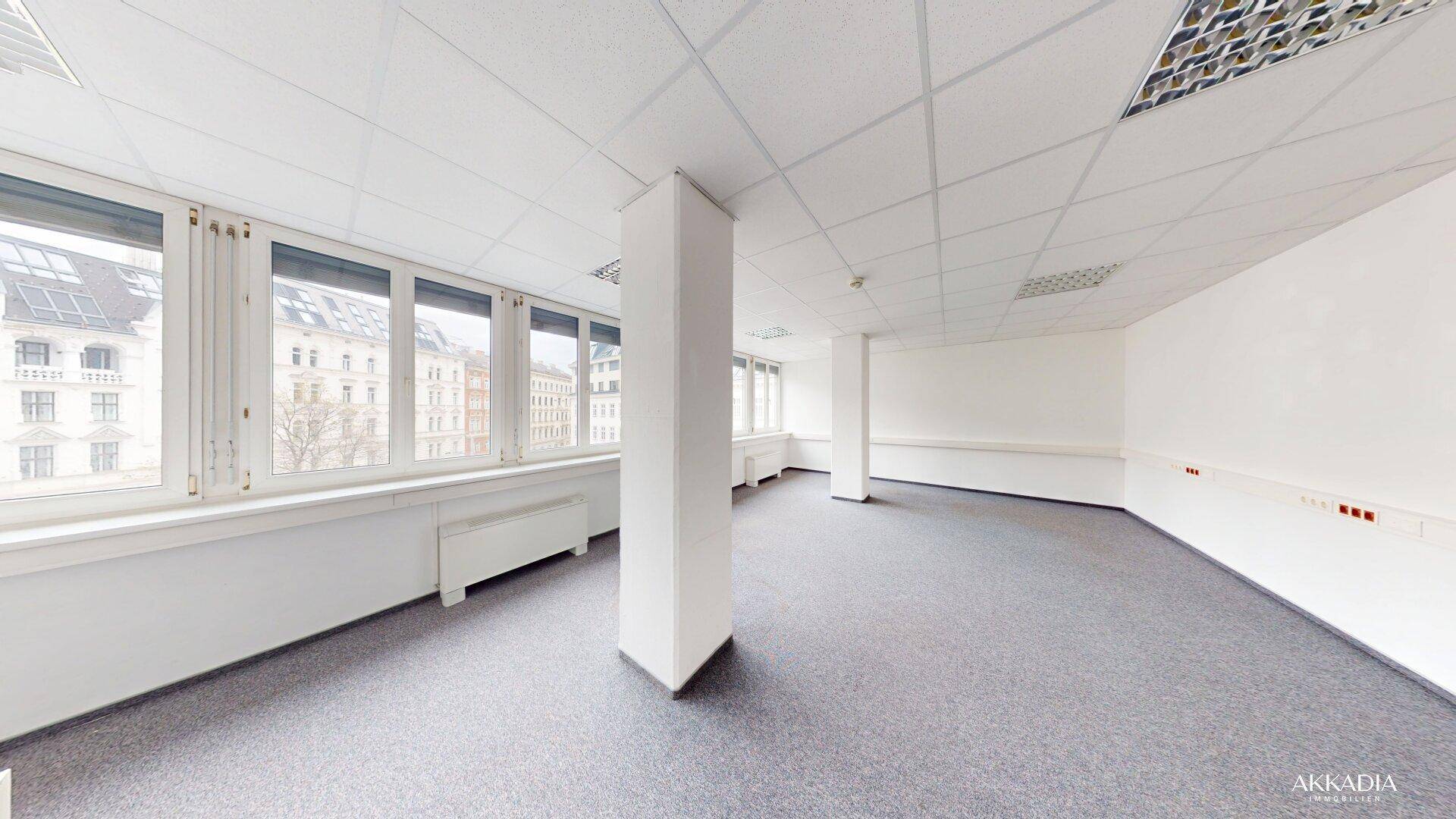Großes Eckbüro mit Teppichboden, Säulen und einer langen Fensterfront mit Stadtblick.