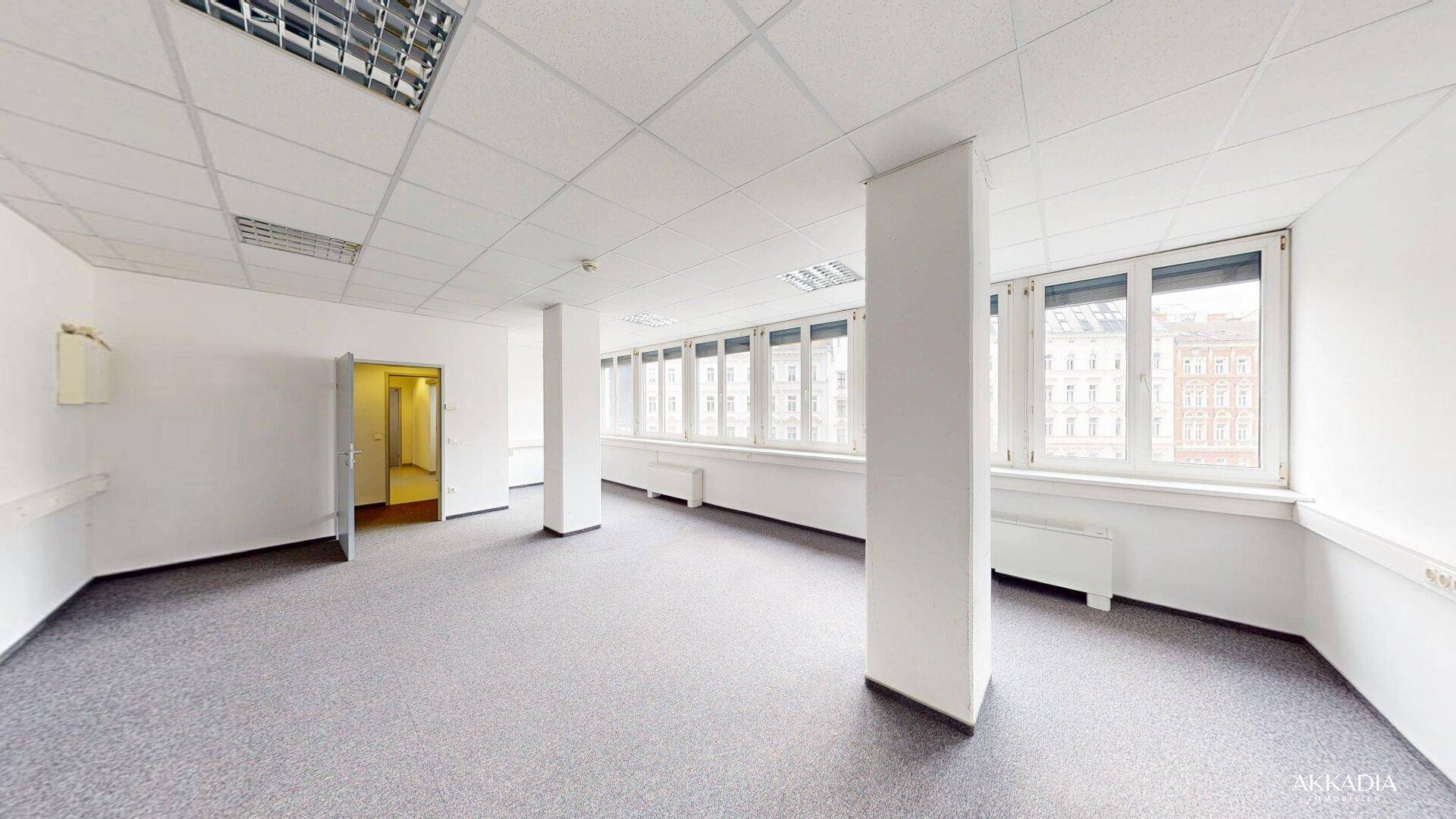 Geräumiges Büro mit Teppichboden, Säulen und Fenstern, die den Raum mit Tageslicht durchfluten.