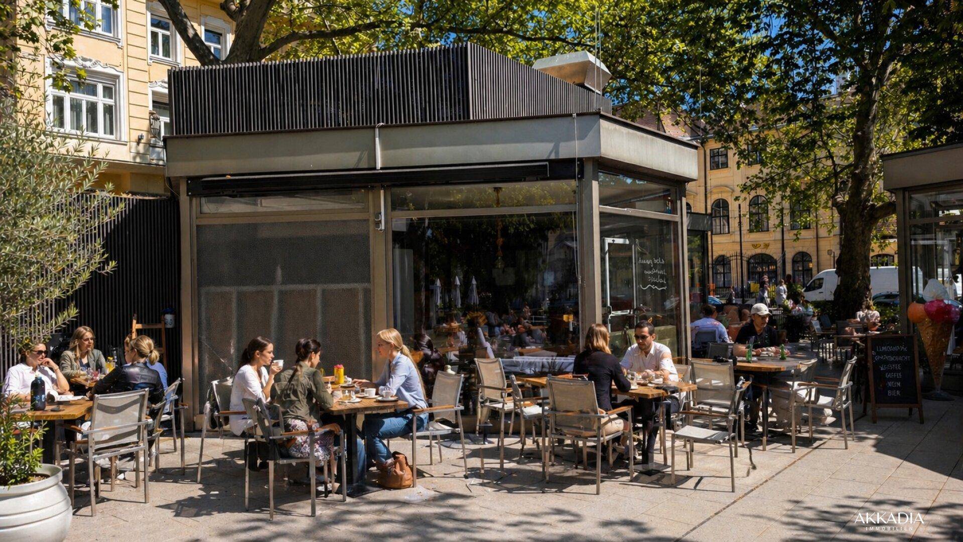 Sonniger Außenbereich eines Cafés mit Tischen und Stühlen, umgeben von Bäumen und Gebäuden.