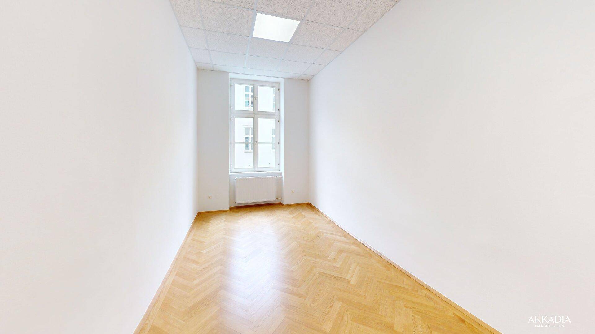 Leerer Raum mit weiß gestrichenen Wänden, Fischgrätparkett und einem großen Fenster.