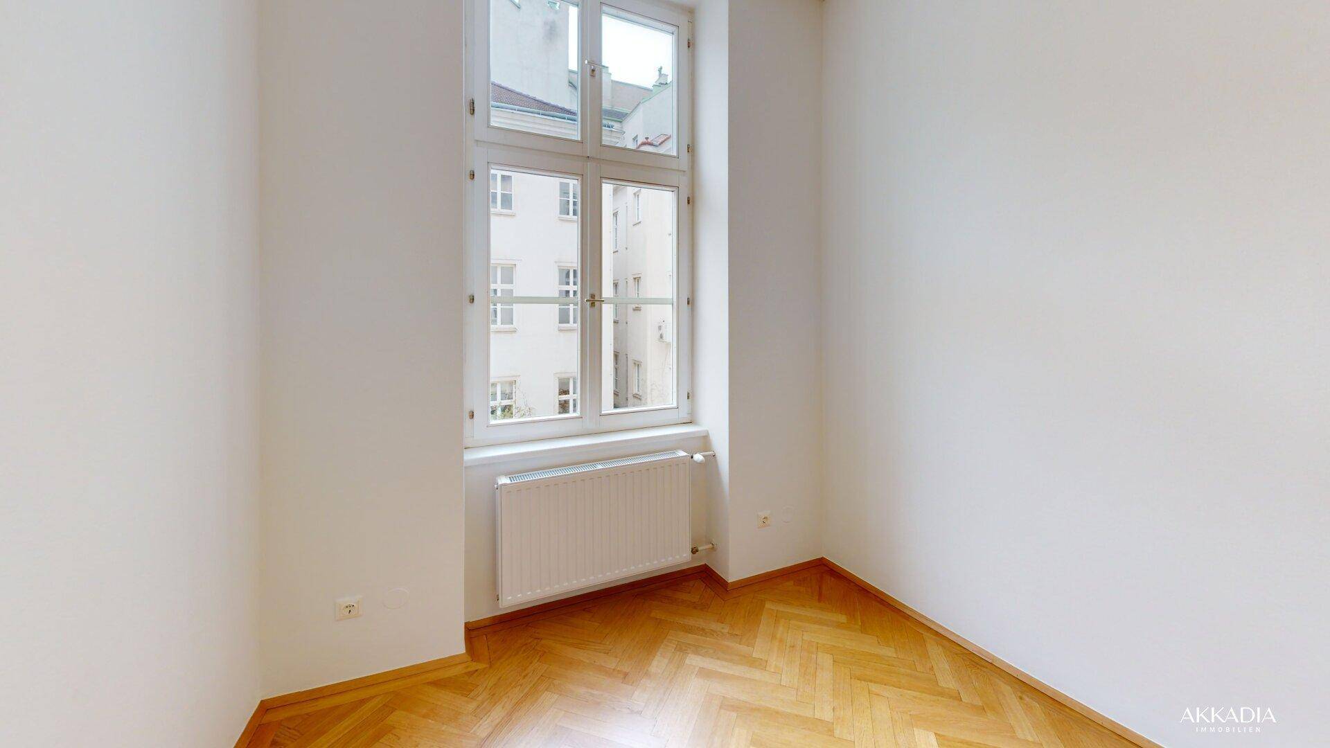 Heller Raum mit Fischgrätparkett und einem hohen Fenster, das auf einen Innenhof blickt.