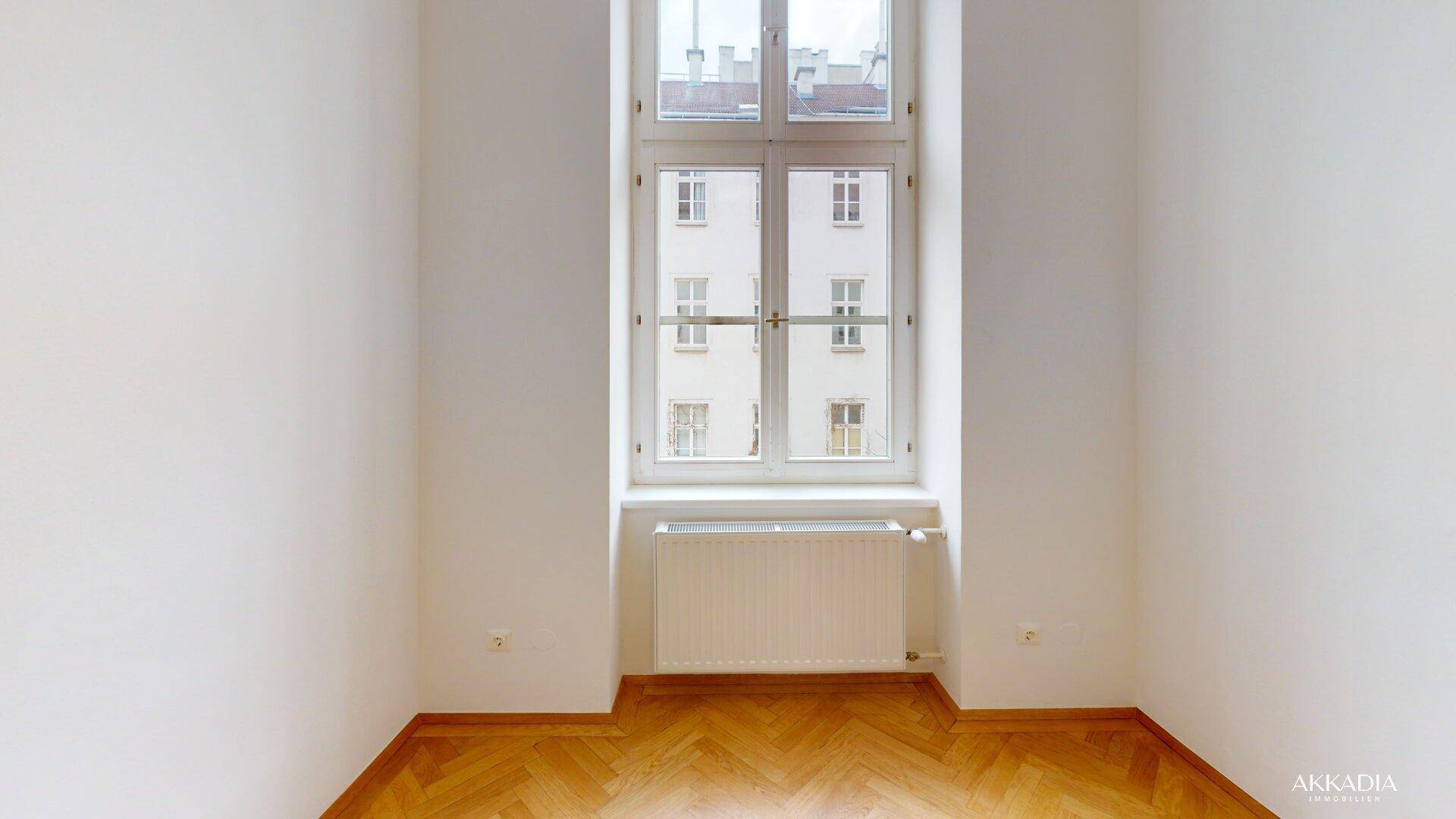 Heller Raum mit Fischgrätparkett und einem hohen Fenster, das auf einen Innenhof blickt.