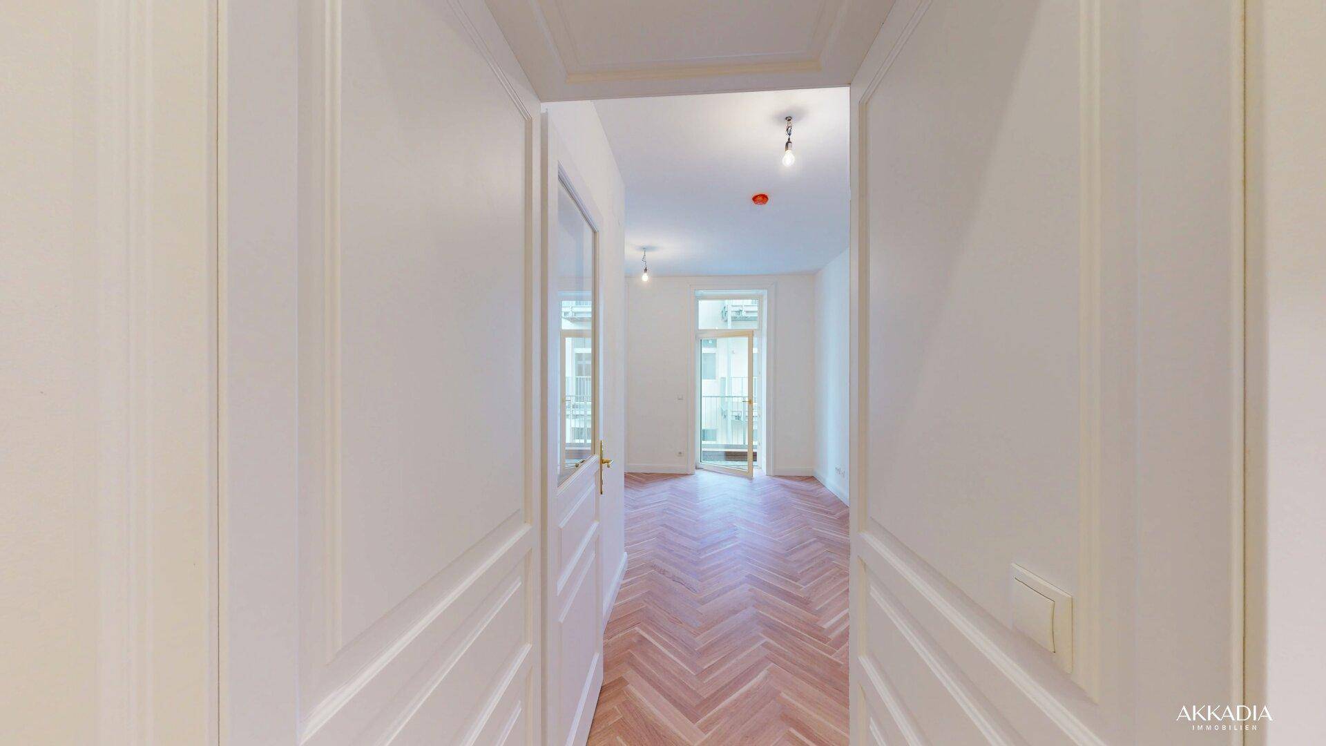 Blick durch eine Tür in einen hellen Raum mit Fischgrätparkett und Zugang zu einem Balkon.