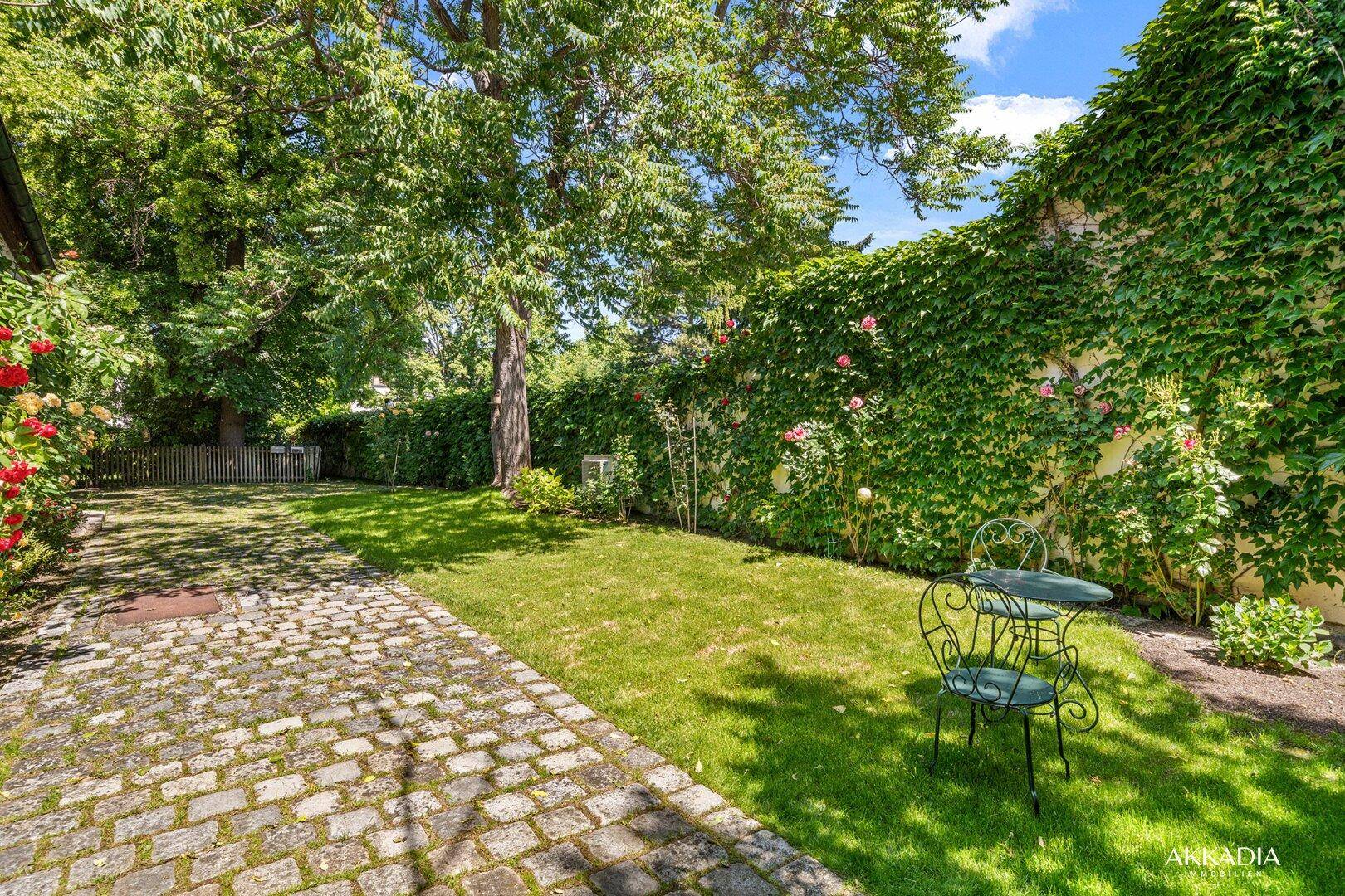 Ruhiger Gartenbereich mit Rasen, gepflastertem Weg und einer kleinen Sitzgruppe unter Bäumen.
