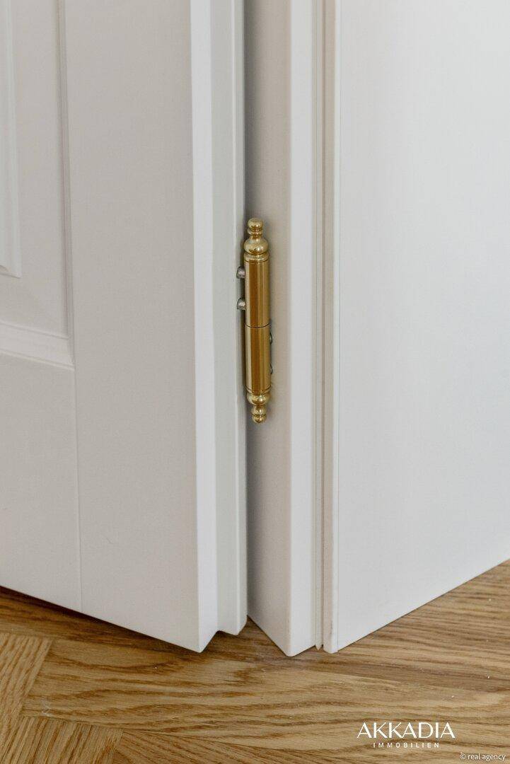 Detailaufnahme eines glänzenden, goldfarbenen Türscharniers und Fischgrätparkettboden.