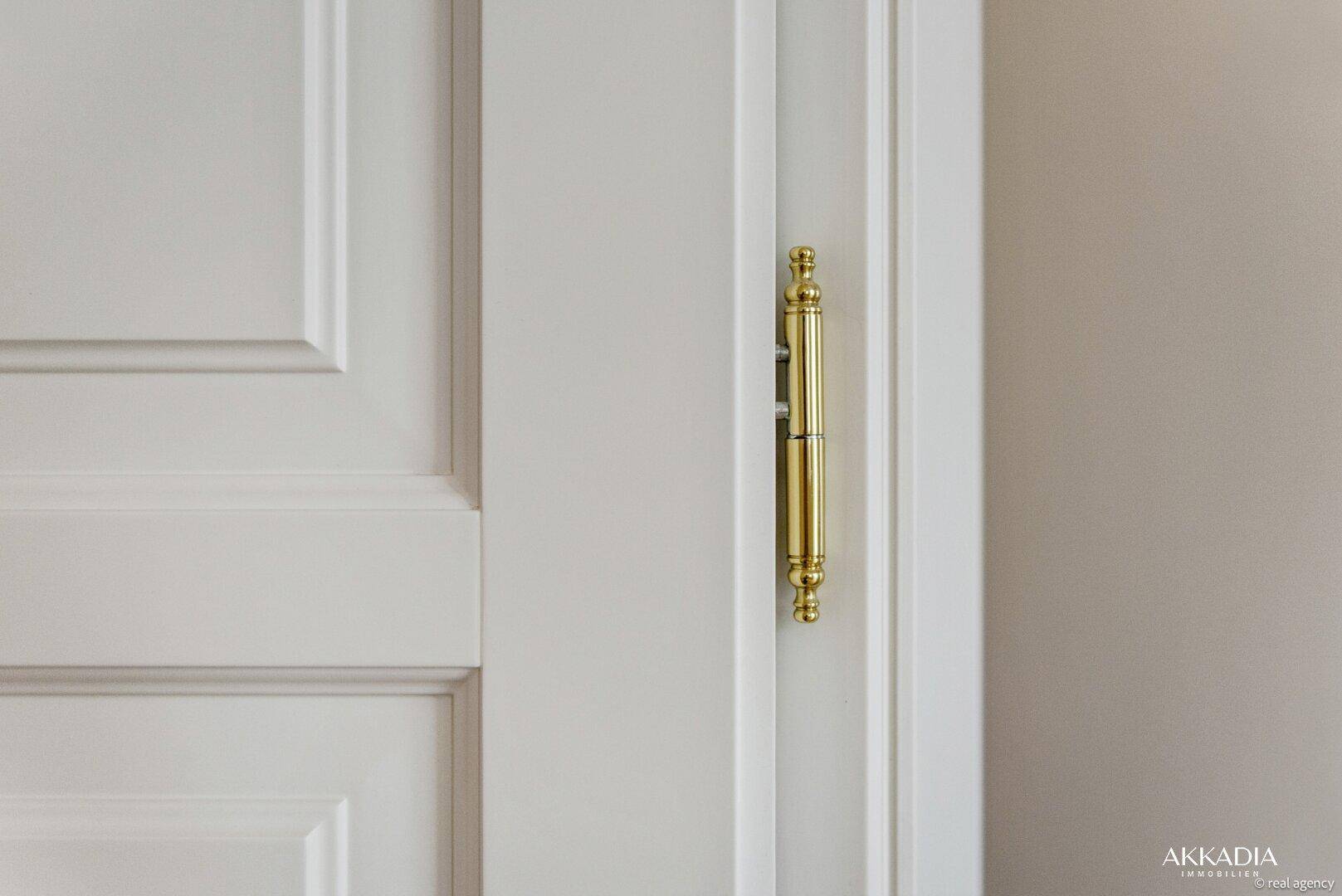 Detailaufnahme eines glänzenden, goldfarbenen Türscharniers an einer weißen Innentür.