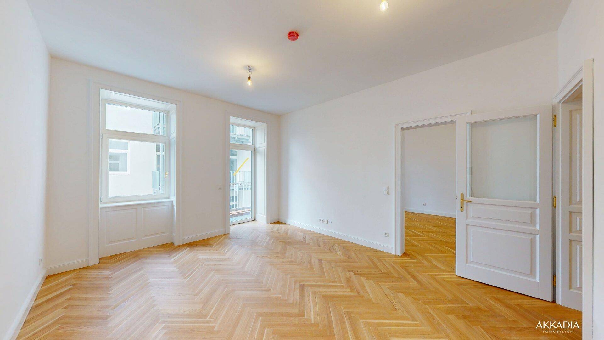 Geräumiges, helles Zimmer mit Fischgrätparkett, großen Fenstern und Zugang zu einem Balkon.