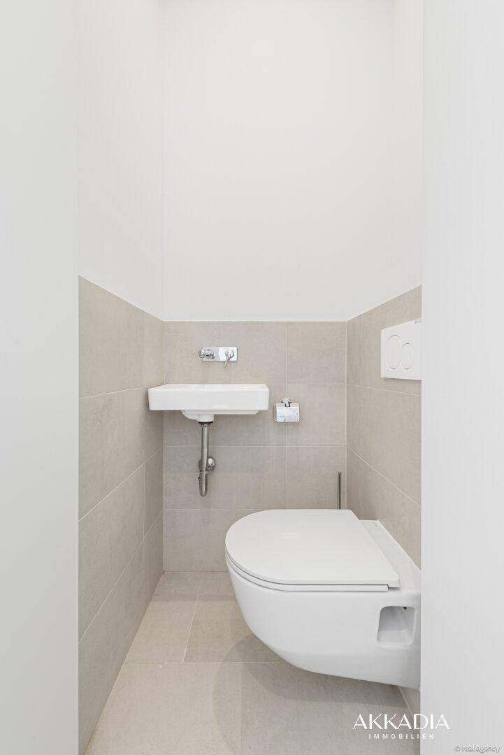 Kompaktes Gäste-WC mit wandhängender Toilette und kleinem Waschbecken, in neutralen Farbtönen gehalten.