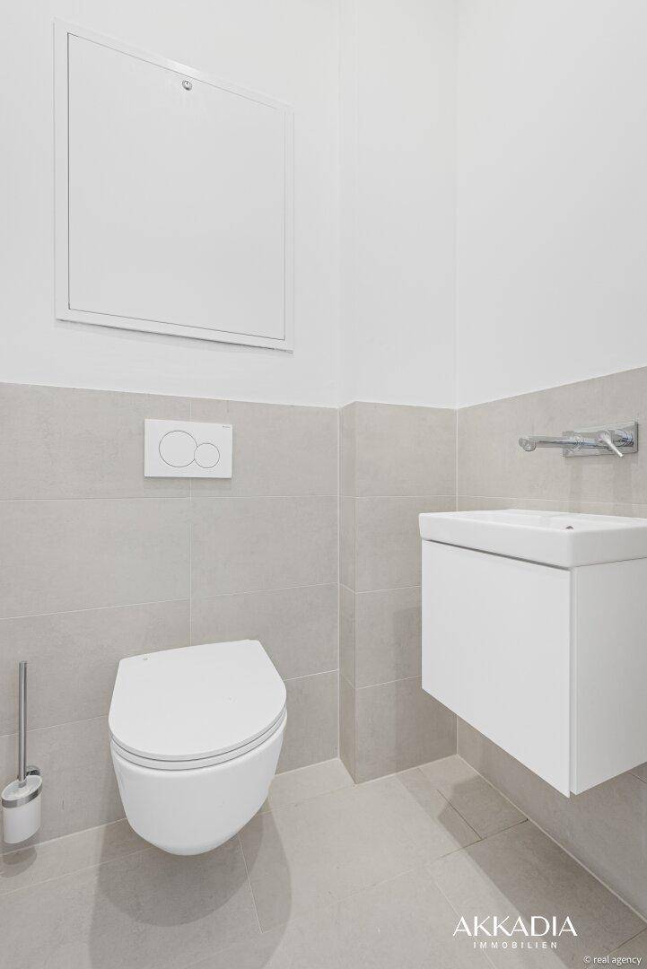 Modernes Gäste-WC mit wandhängender Toilette und kleinem Waschbecken in hellgrauen Fliesen.