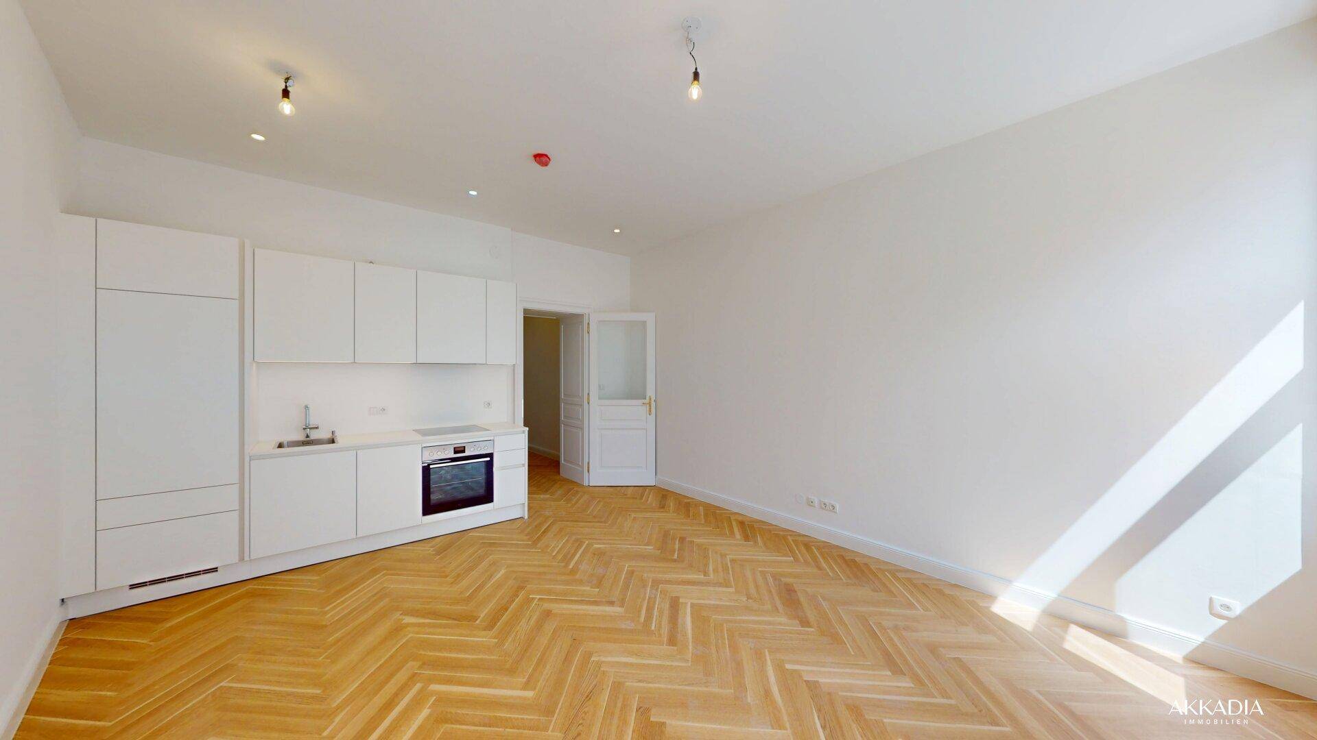 Offener Wohnbereich mit moderner weißer Einbauküche, integriertem Backofen und elegantem Fischgrätparkett.