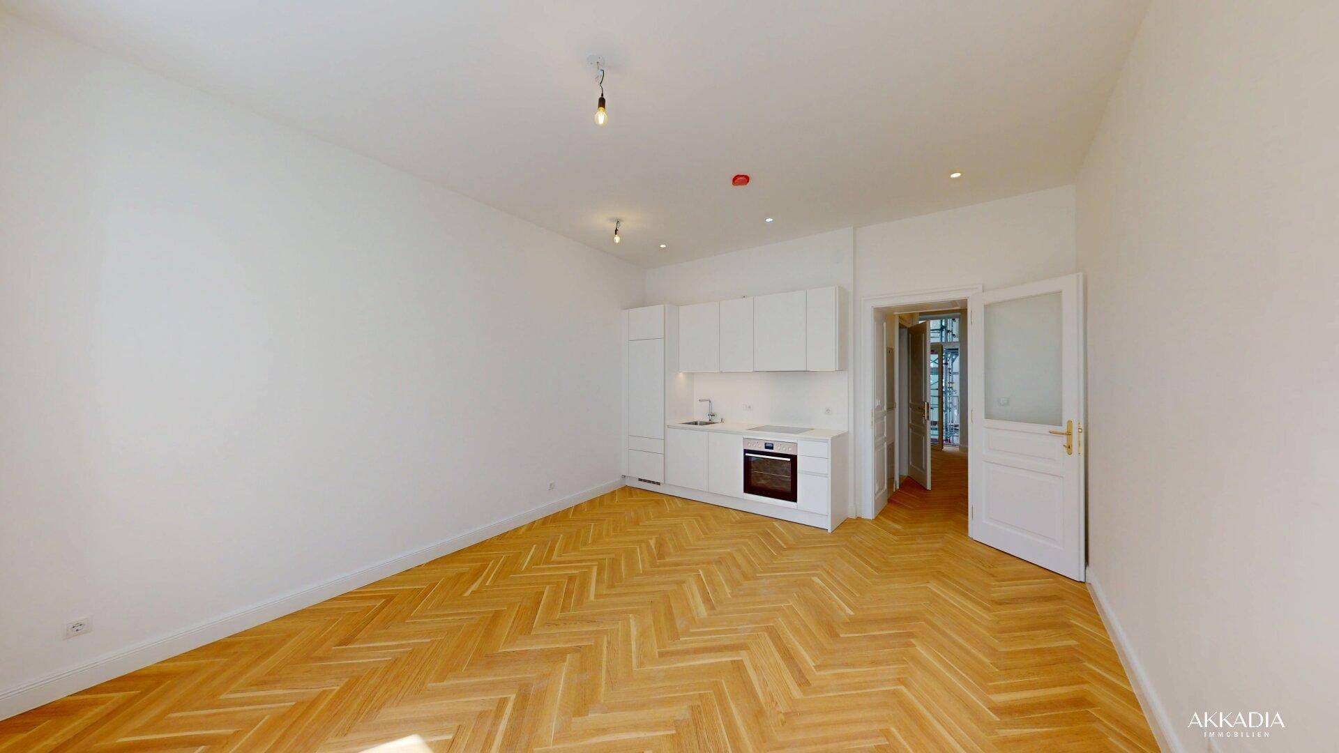 Geräumiger Wohnbereich mit moderner Einbauküche, Fischgrätparkett und Zugang zu einem weiteren Zimmer.