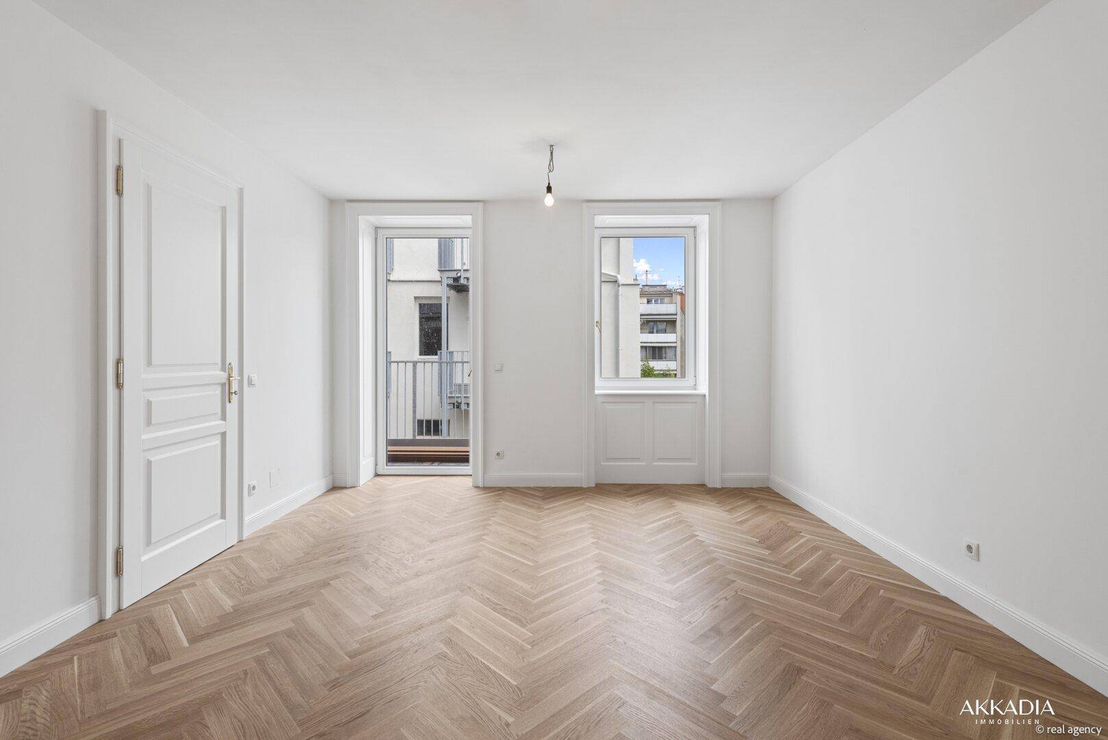 Geräumiges Wohnzimmer mit Fischgrätparkettboden und Zugang zum Balkon.