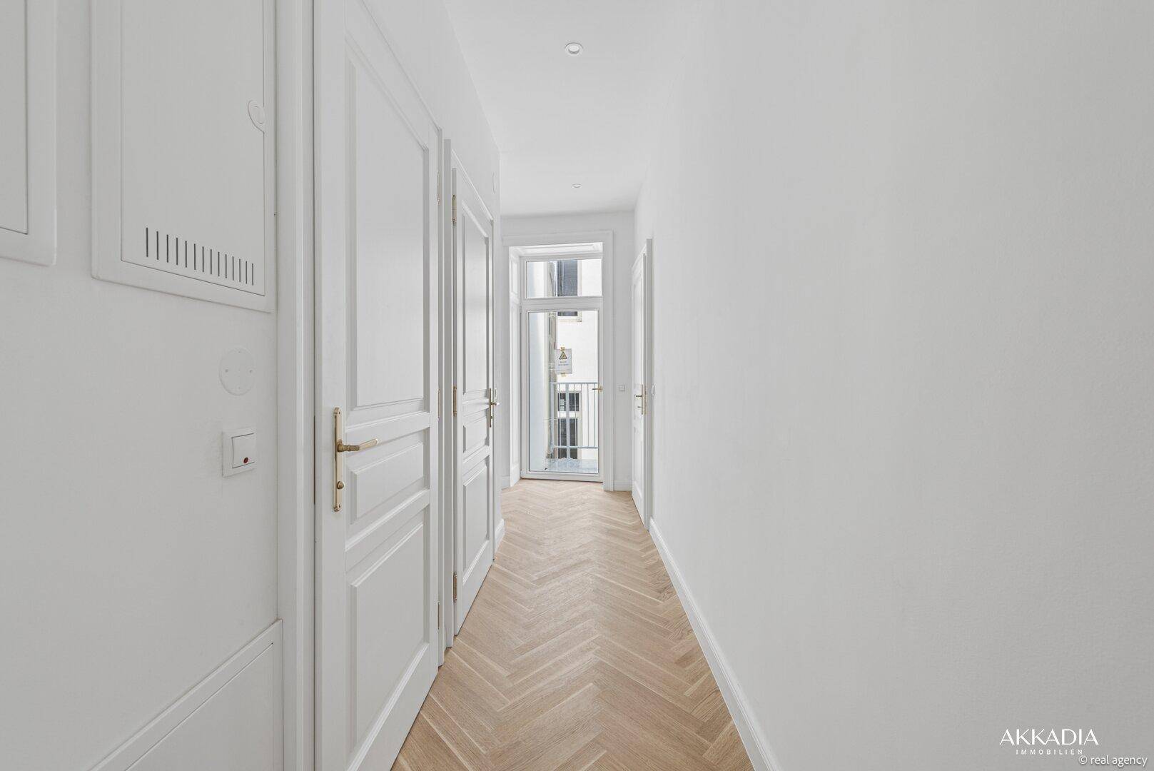 Heller Flur mit Fischgrätparkett, weißen Türen und Zugang zu einem Balkon.