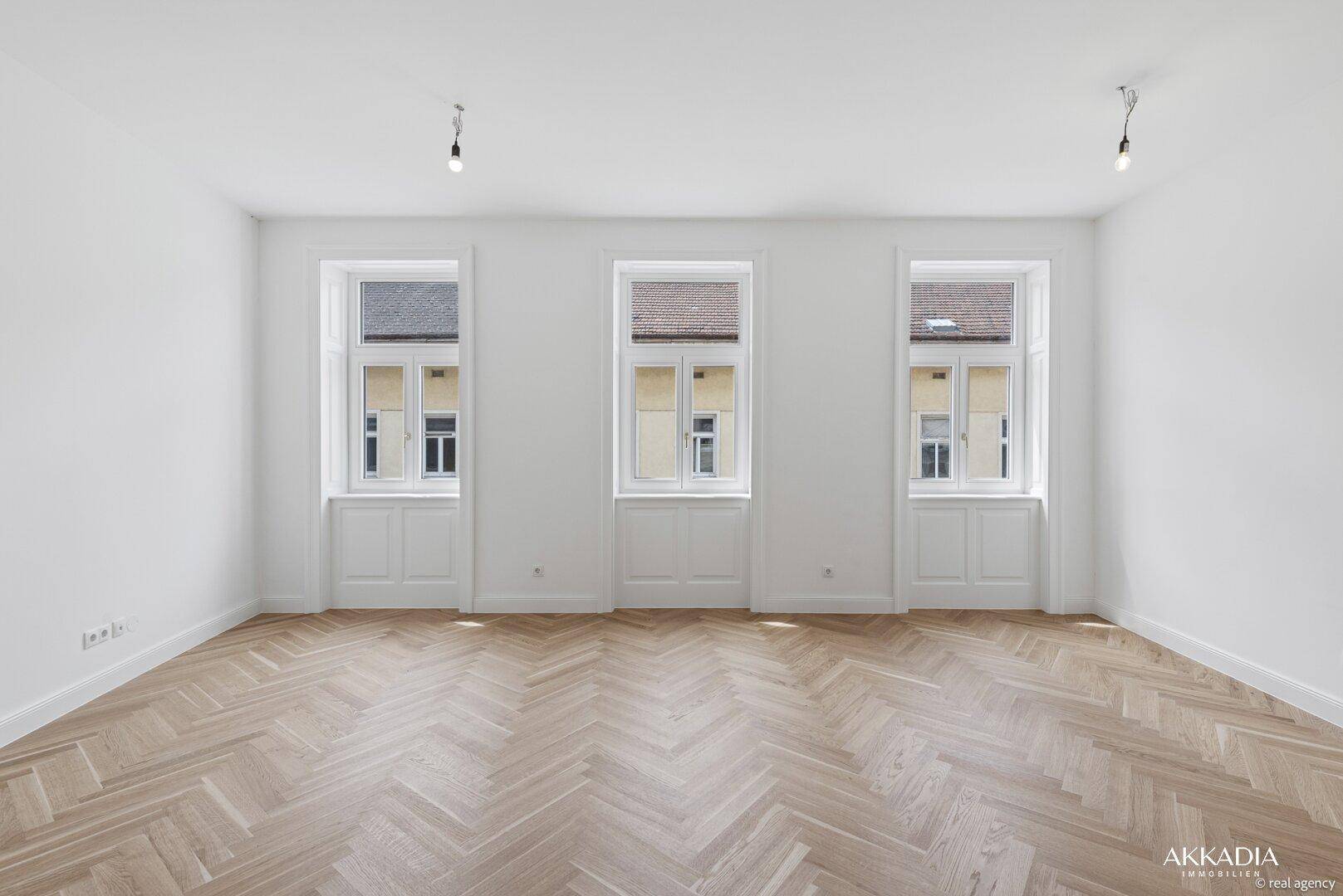 Geräumiges, helles Zimmer mit Fischgrätparkett und drei hohen Fenstern, ideal als Wohnzimmer.