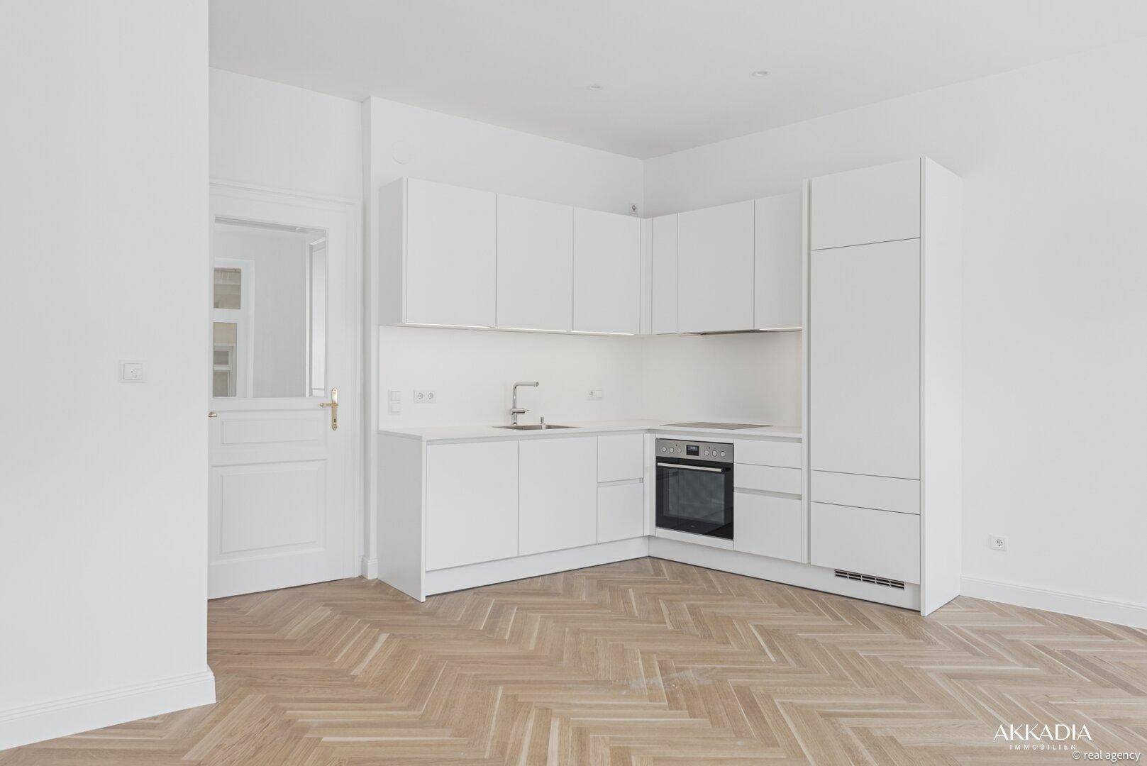 Moderne, weiße Einbauküche mit integrierten Geräten und Fischgrätparkett in einem hellen Raum.
