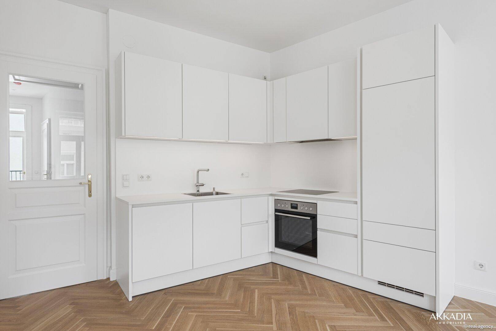 Moderne, weiße Einbauküche mit integrierten Geräten und Fischgrätparkett in einem hellen Raum.