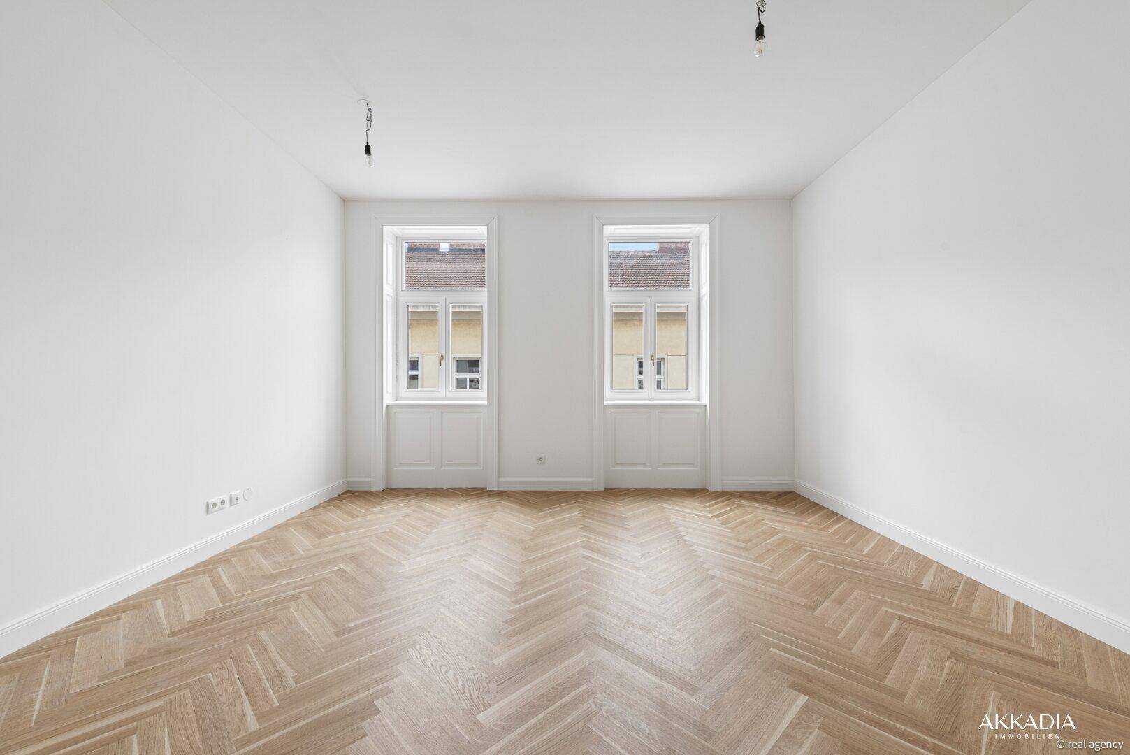 Geräumiger, heller Raum mit elegantem Fischgrätparkett und zwei großen Fenstern für viel Tageslicht.