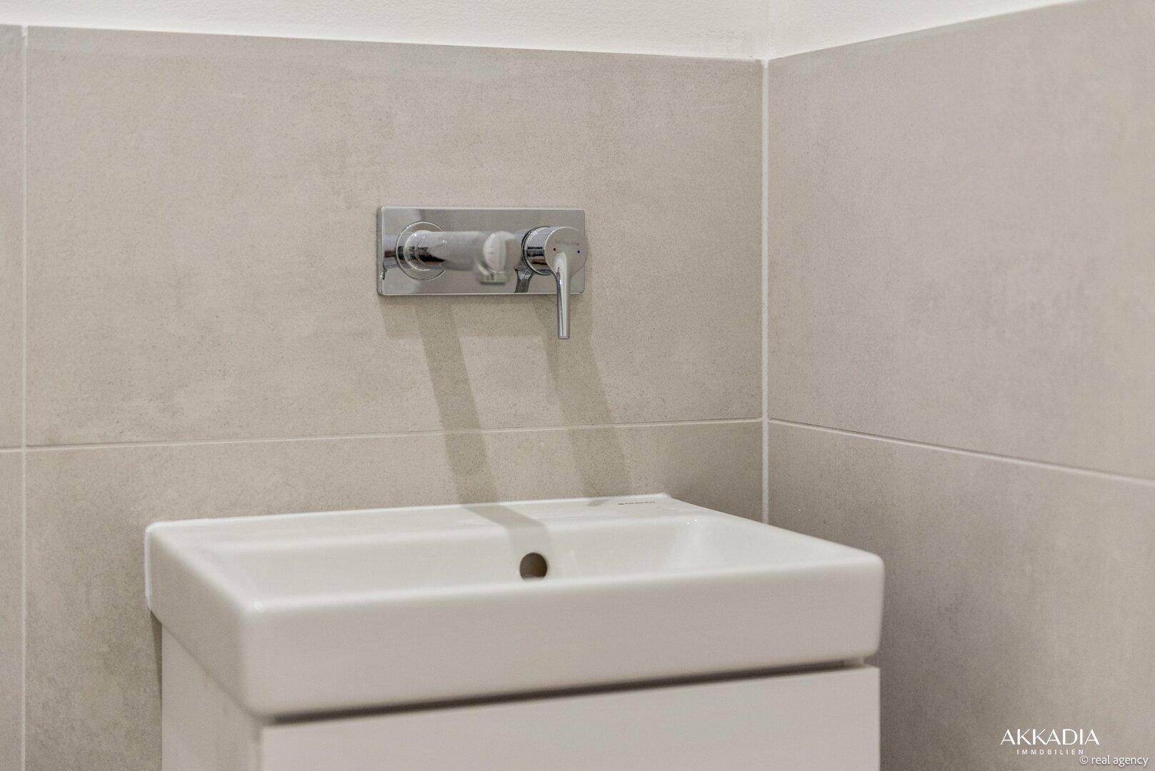 Kleines Waschbecken mit modernem Wandwasserhahn in einem hell gefliesten Gäste-WC.