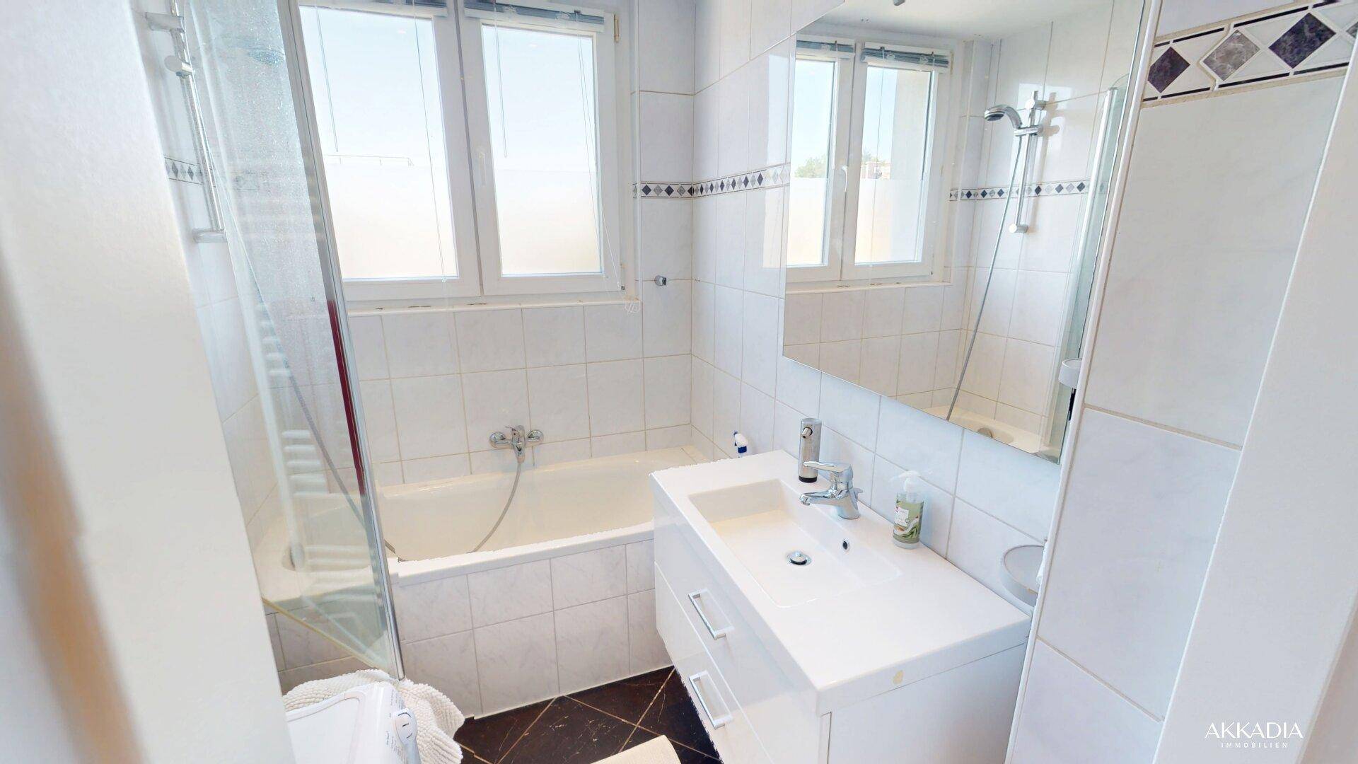 Helles Badezimmer mit weißen Fliesen, Badewanne mit Duschfunktion und einem Fenster für Tageslicht.