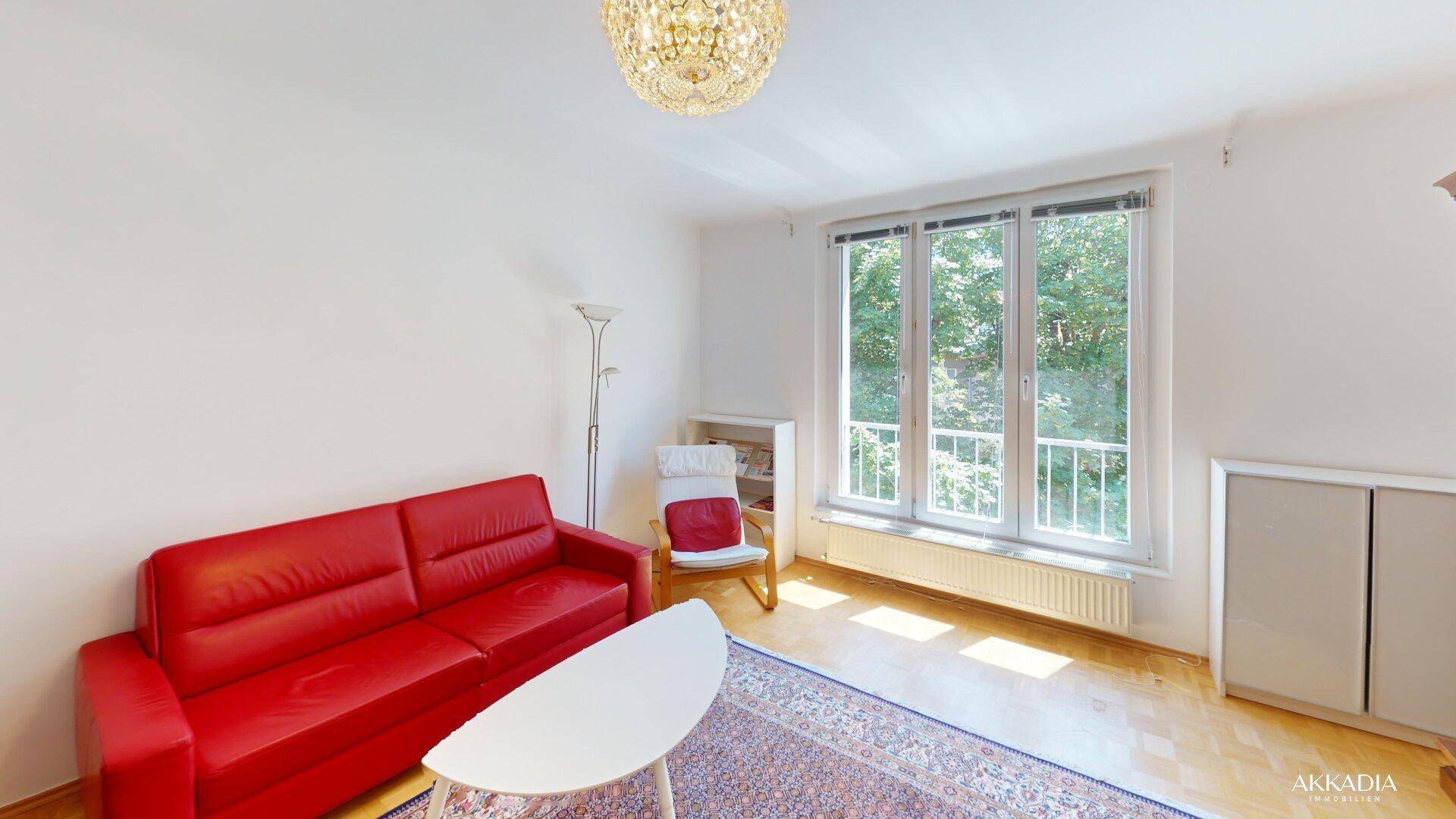 Geräumiges Wohnzimmer mit rotem Sofa, Parkettboden und einem Fenster mit Blick ins Grüne.