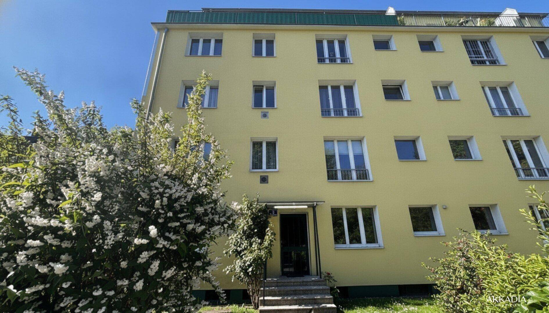 Gelbe Fassade eines Mehrfamilienhauses mit vielen Fenstern und blühenden Sträuchern im Vordergrund.