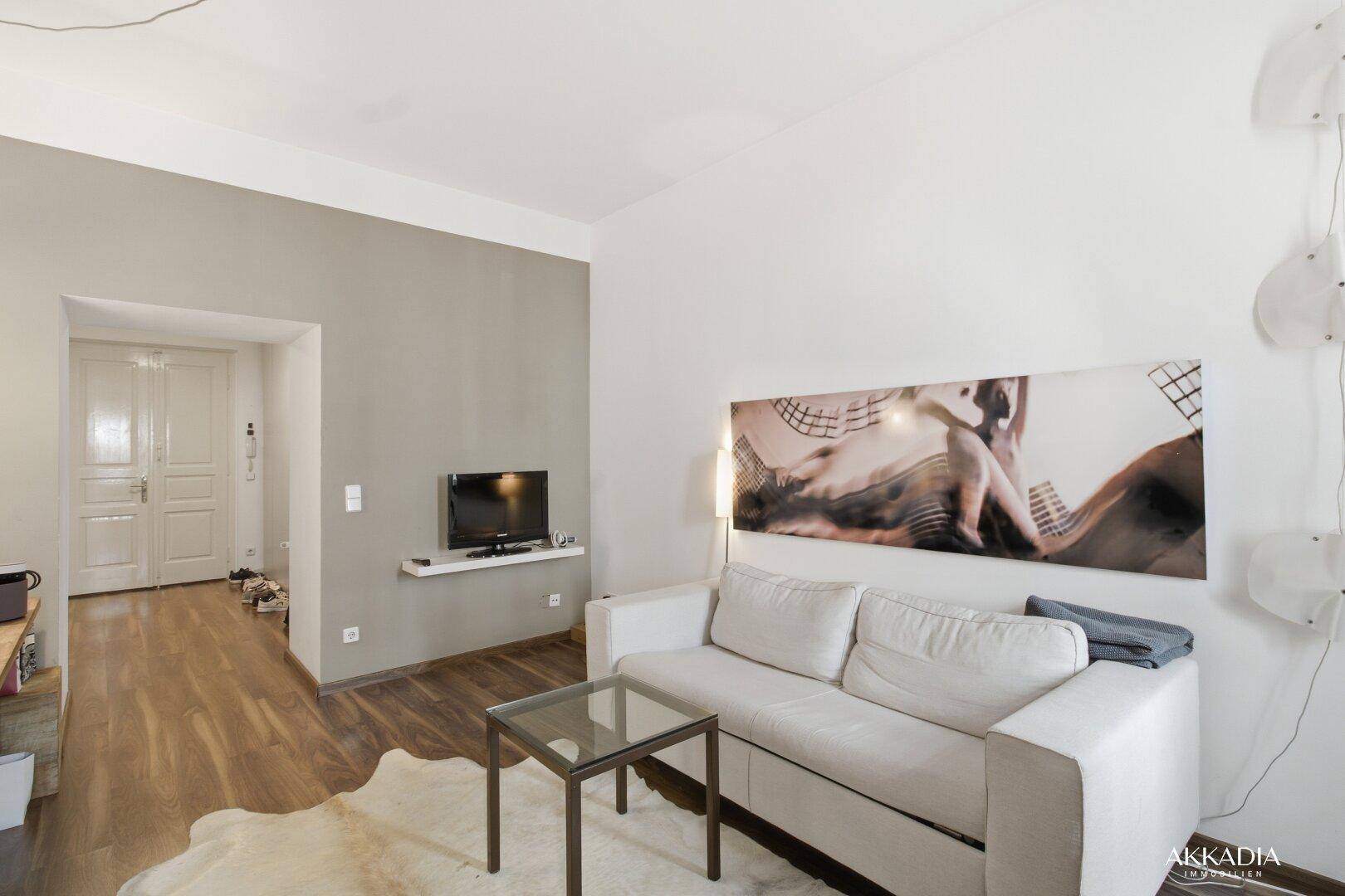 Wohnzimmerbereich mit Fernseher, hellem Sofa und Zugang zu einem weiteren Raum.