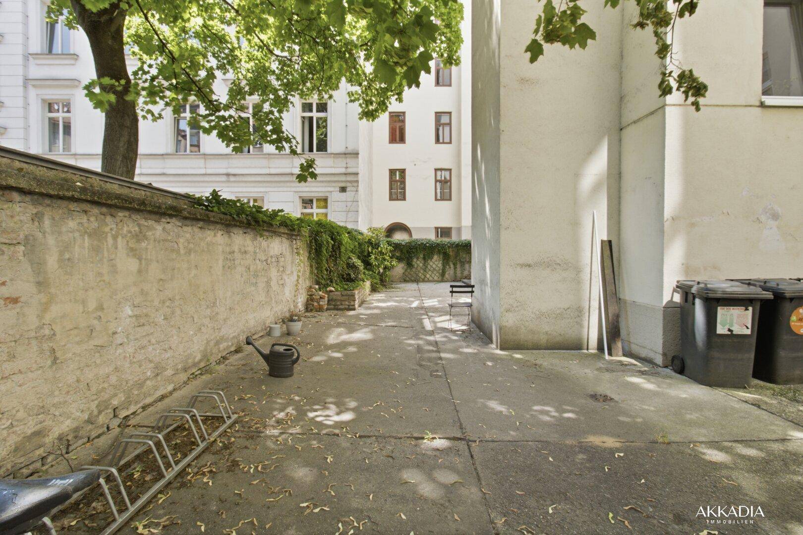 Gepflasterter Hinterhofbereich mit Grünpflanzen und Mülltonnen.
