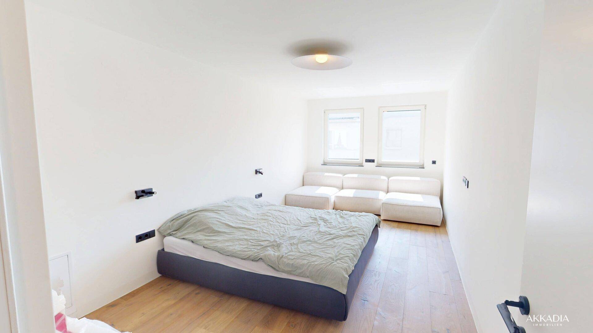 Helles Schlafzimmer mit weiß gestrichenen Wänden, Holzboden und zwei Fenstern für natürliche Belichtung.