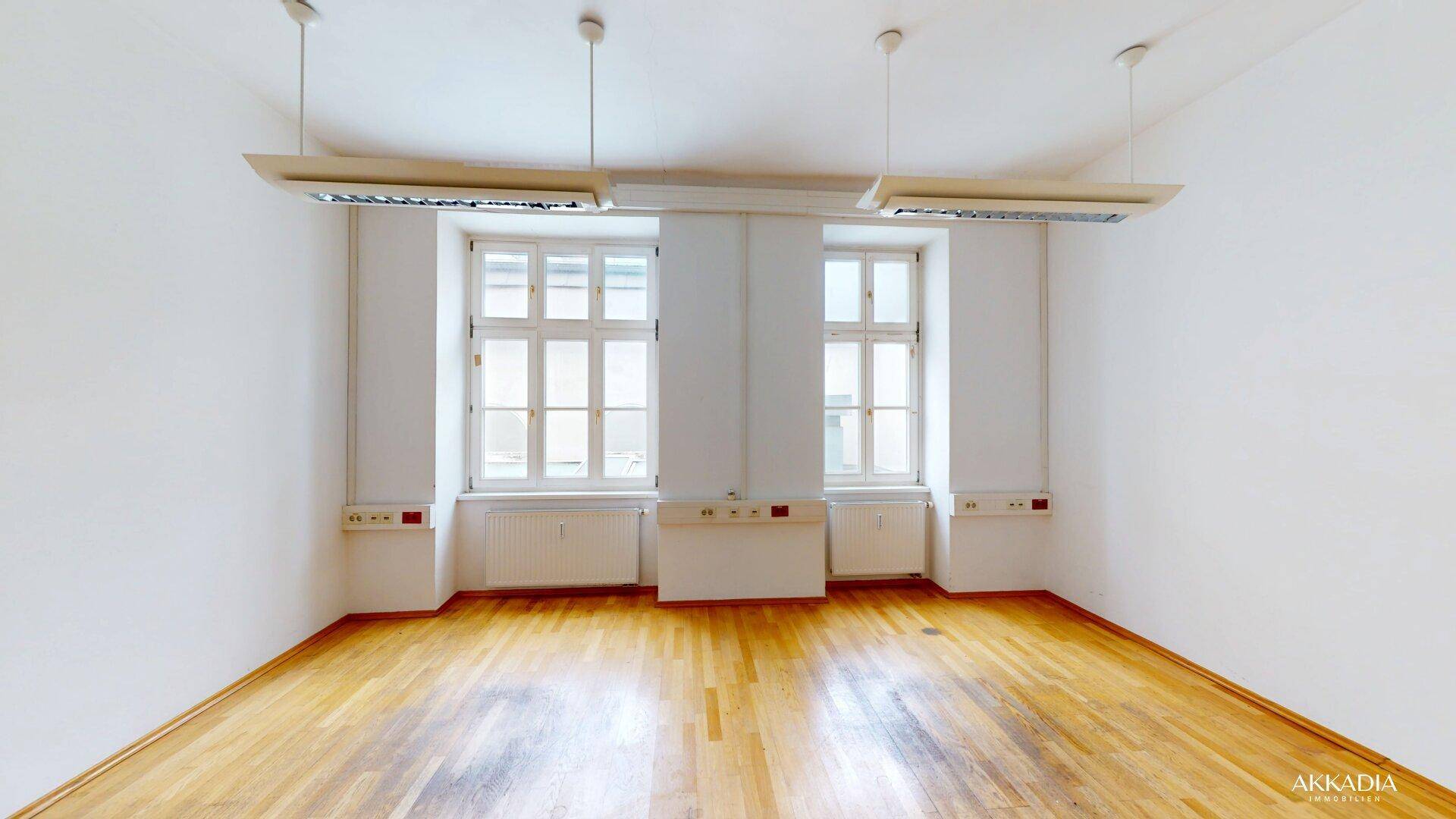 Heller Büroraum mit zwei großen Fenstern, Holzfußboden und modernen Deckenleuchten für eine gute Ausleuchtung.