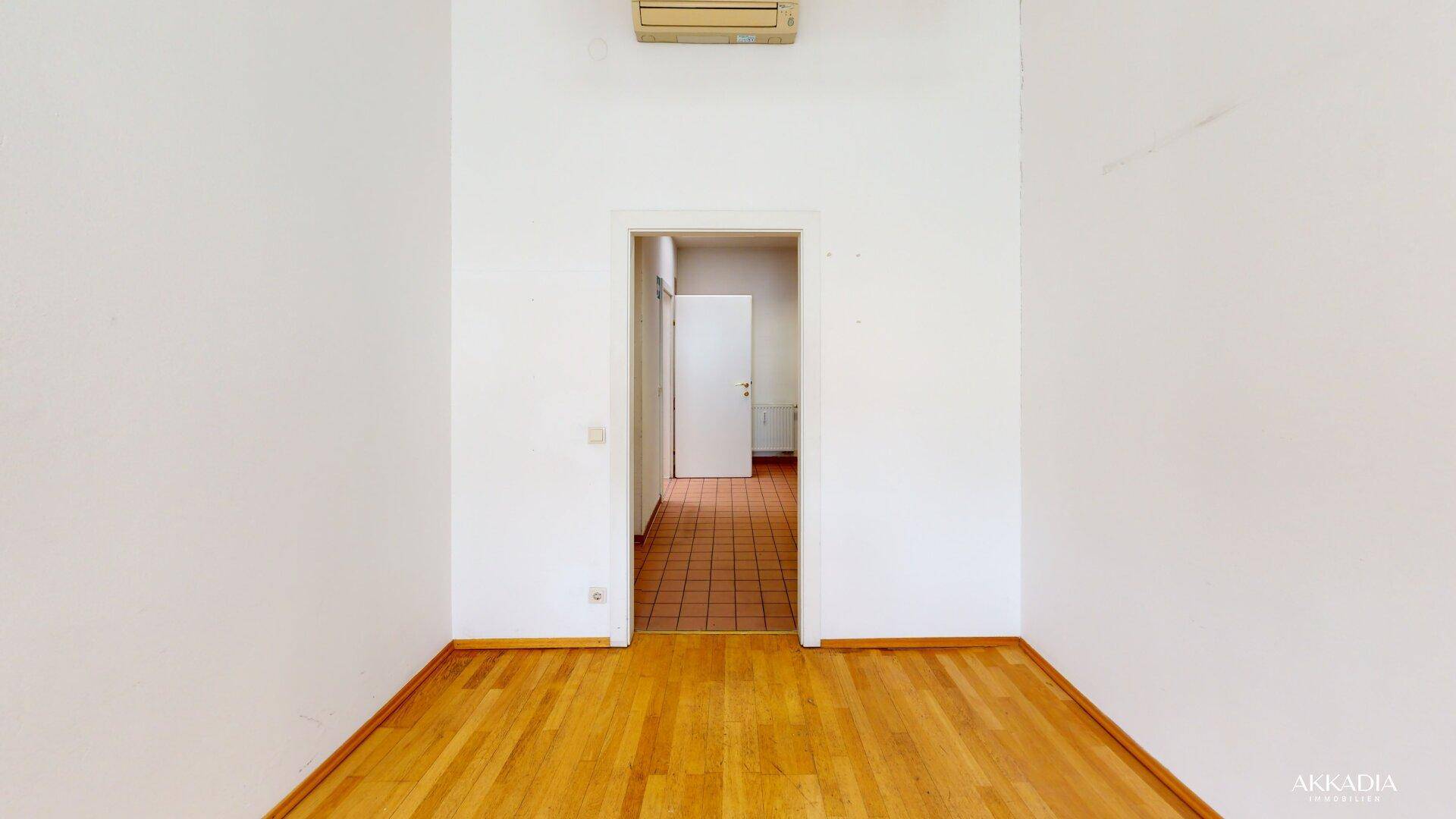 Büroraum mit weiß gestrichenen Wänden, Holzfußboden und einer Tür, die zu einem gefliesten Bereich führt.