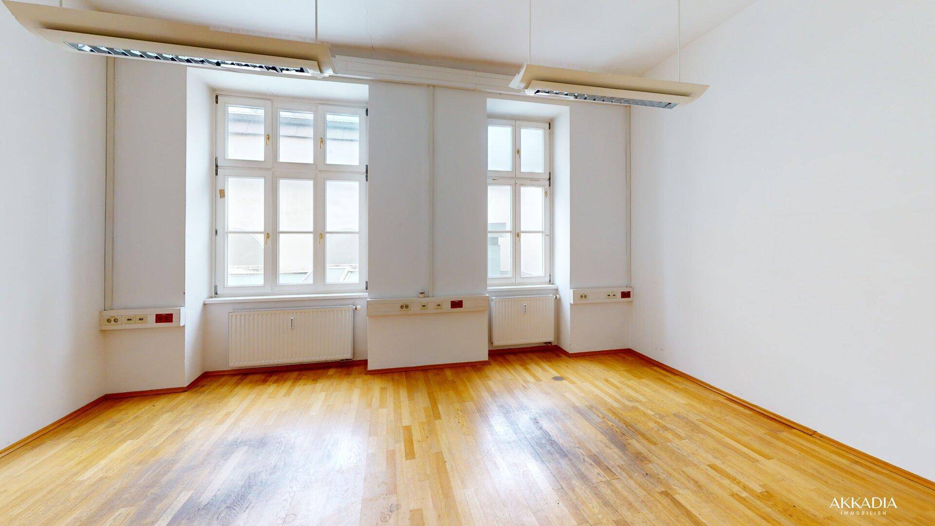 Heller Büroraum mit zwei großen Fenstern, Holzfußboden und modernen Deckenleuchten für eine gute Ausleuchtung.