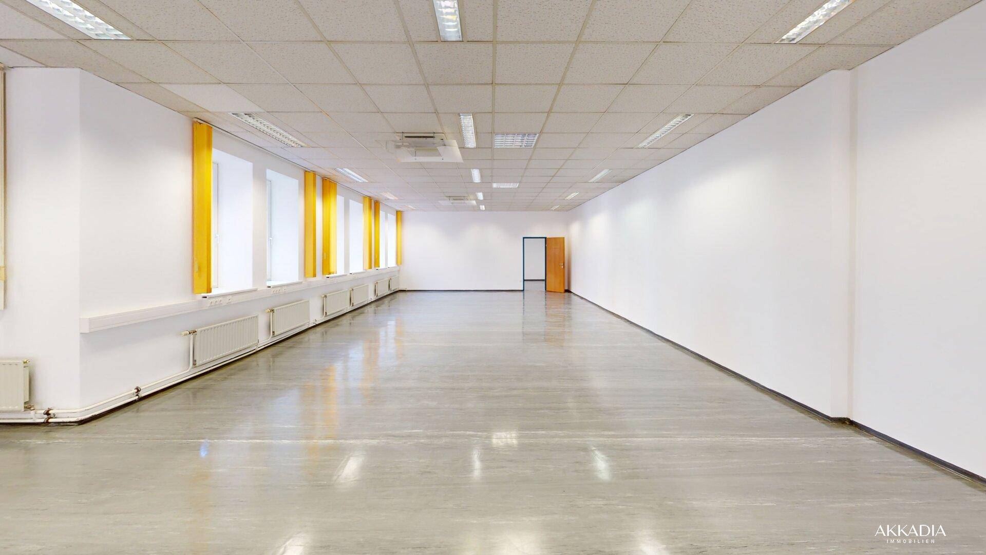 Langer, heller Büroraum mit weißen Wänden, grauem Boden und gelben Jalousien an den Fenstern.