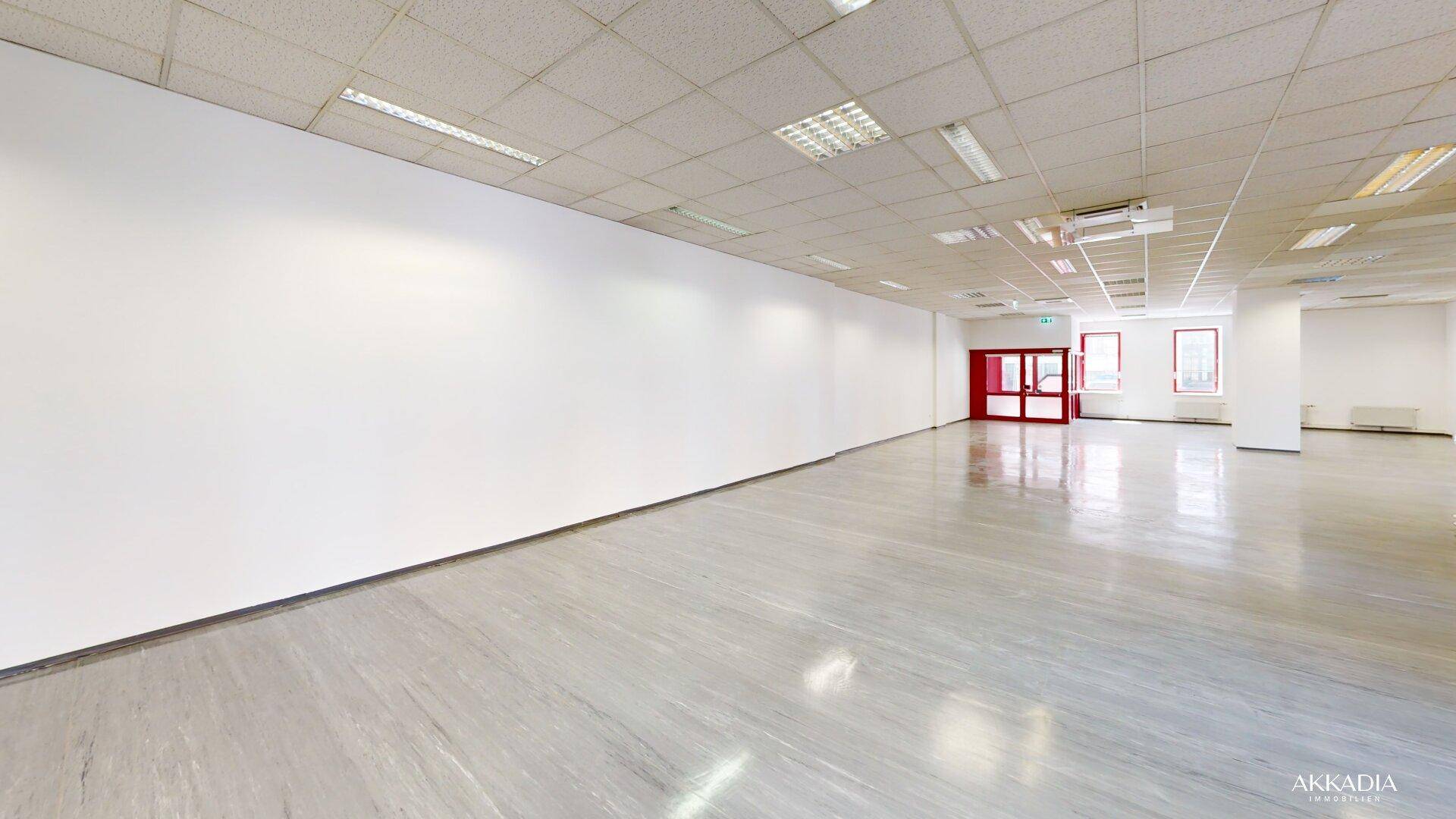 Großer, offener Bürobereich mit weißen Wänden, glänzendem Boden und roten Türen im hinteren Bereich.