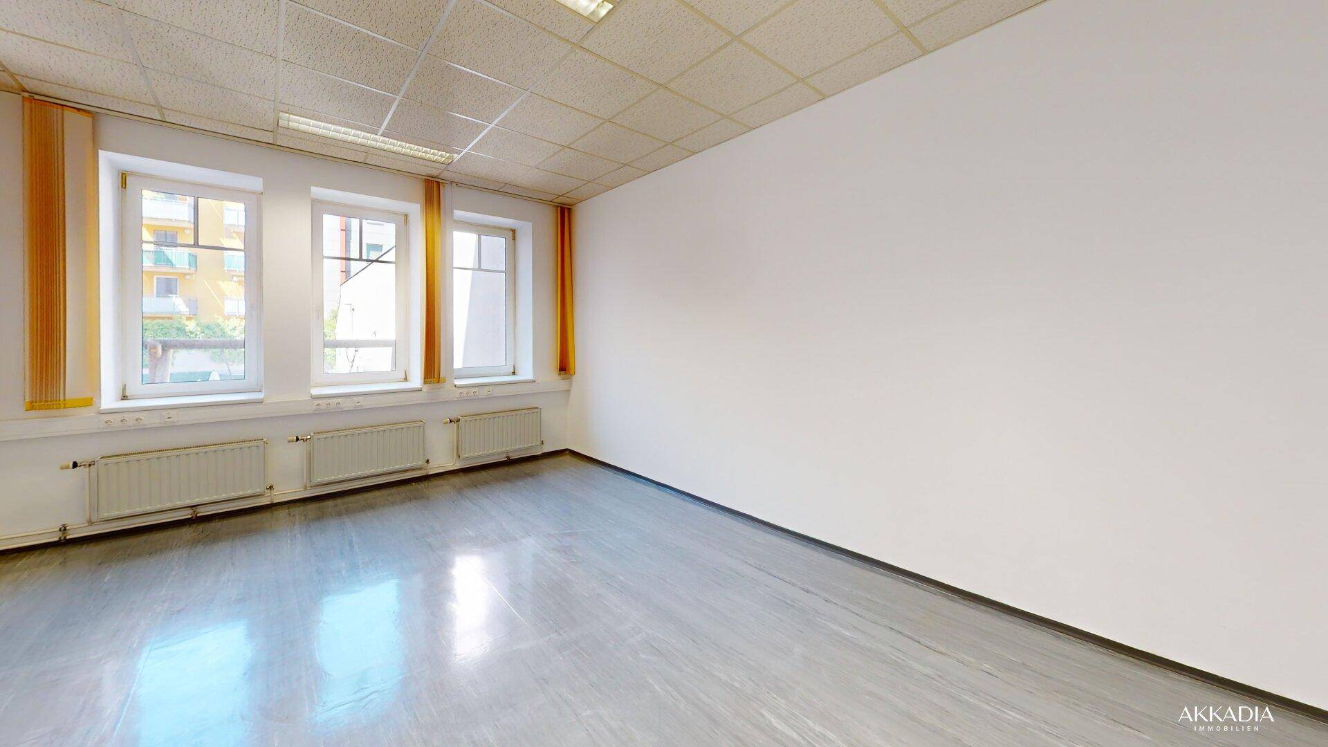 Leerer Büroraum mit weißen Wänden, grauem Boden und drei Fenstern mit Blick ins Grüne.
