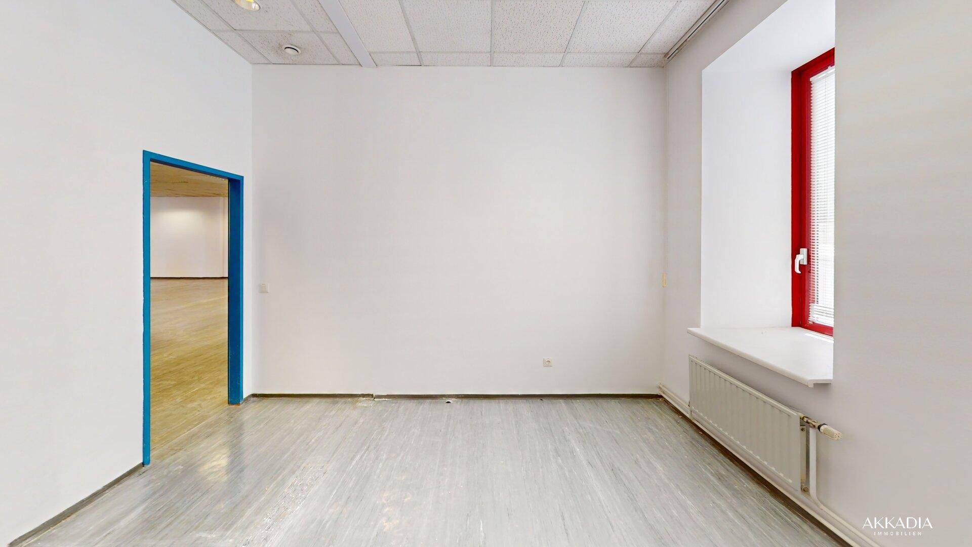 Heller Büroraum mit weißen Wänden, grauem Boden und einem roten Fenster sowie einer blauen Tür.